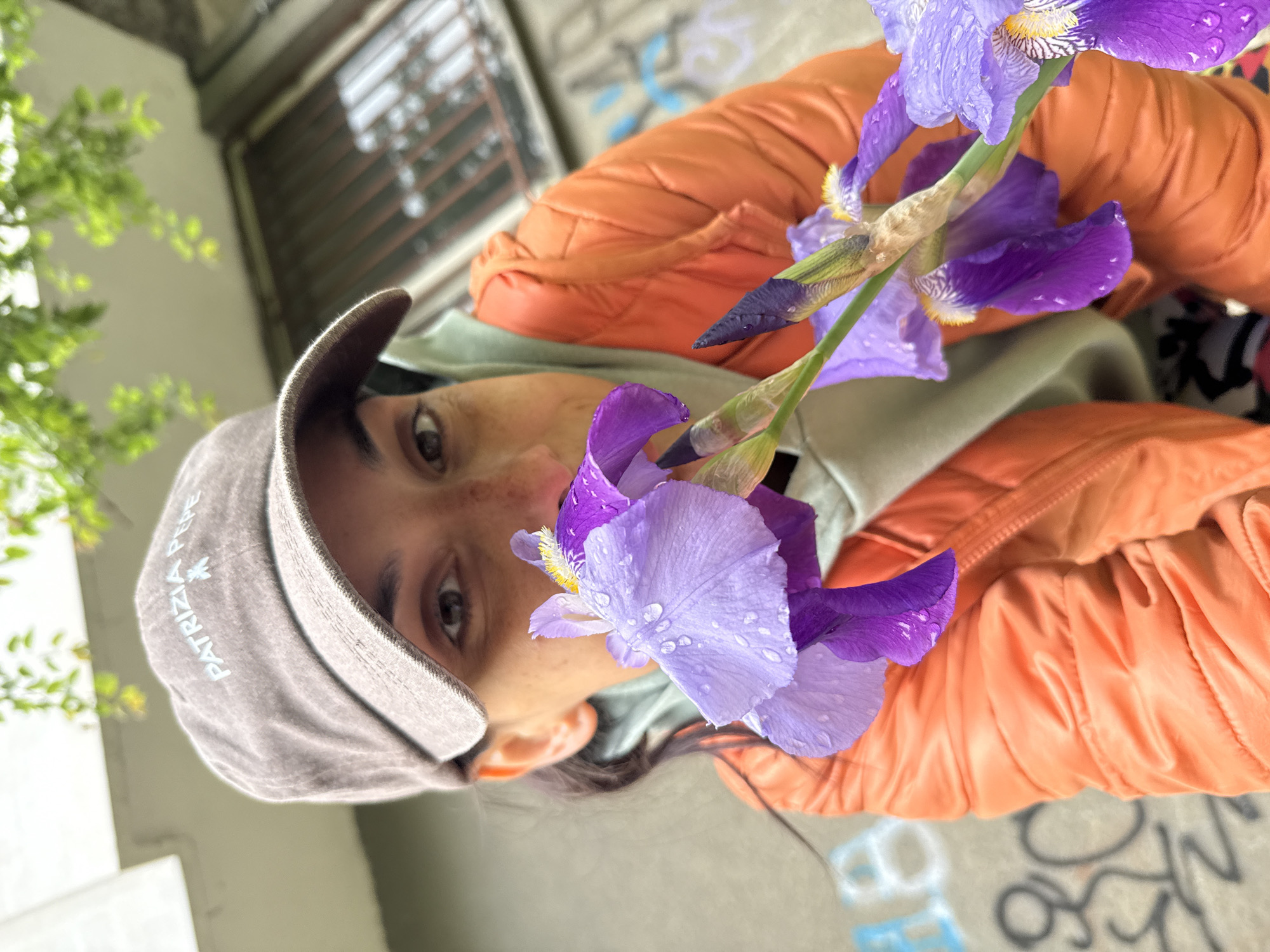 A woman in an orange jacket and a Patrizia Pepe cap holds a bouquet of purple irises close to her face. The background includes a building with a barred window, green foliage, and graffiti-covered concrete. The image suggests an outdoor location. There is no confirmation of it being in Pleven, Bulgaria.
