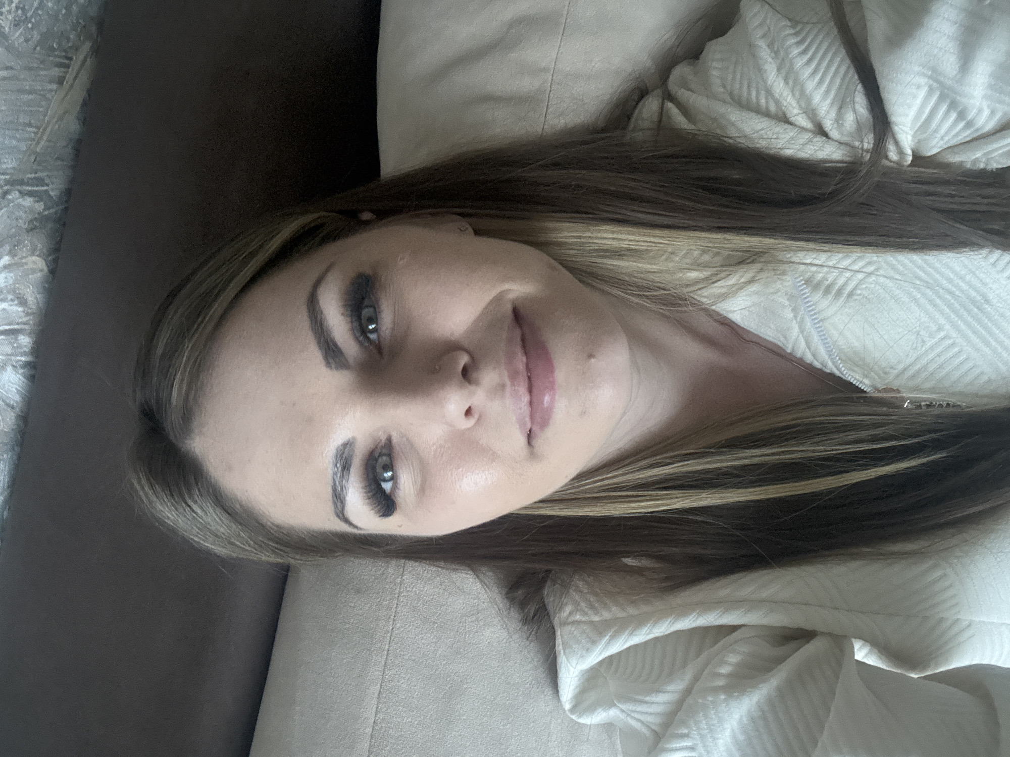 A person is lying down, seemingly on a beige upholstered sofa. They are fair-skinned with long, brown hair featuring blonde highlights. Their eyes are visible, enhanced with dark eye makeup, including distinct eyelashes. They wear what appears to be a light-colored, quilted garment. Part of a brown upholstered surface is visible on the left edge of the frame. The image's orientation is sideways. There is no definitive indication of the location being in the city of Razdolie, Russia.