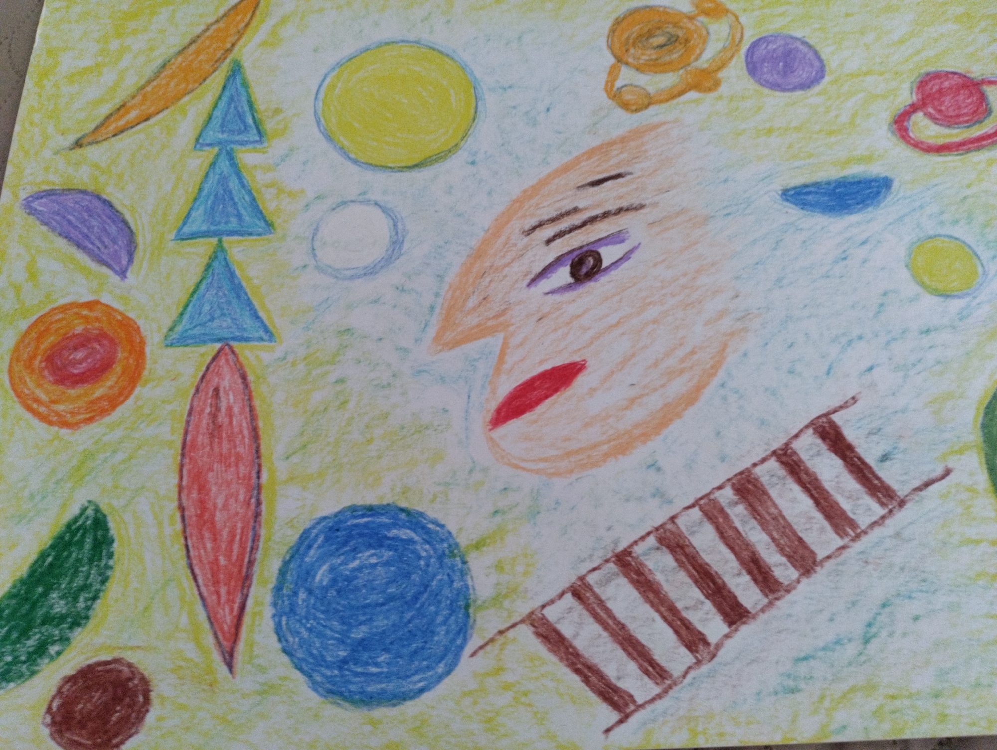 This is a close-up, eye-level, medium shot of a crayon drawing on a light yellow and green textured paper. The drawing depicts an abstract composition with a stylized human face on the right side. The face has an orange skin tone, a prominent nose, a purple eye with dark pupils, and red lips. Above the face, there are several abstract shapes, including yellow circles, purple circles, and orange ring-like forms. To the left of the face, there is a vertical arrangement of three blue triangles, with a long, thin red shape below them. Other shapes in the drawing include a purple crescent, a green elongated oval, a red-orange circle with concentric rings, a brown oval, and a large blue circle. In the lower right portion of the drawing, there is a diagonal representation of a striped structure, possibly a ladder or stairs, with brown stripes. The drawing is rendered in a child-like, abstract style. There is no indication of the city of Mezitli, Turkey, or any real-world environment.