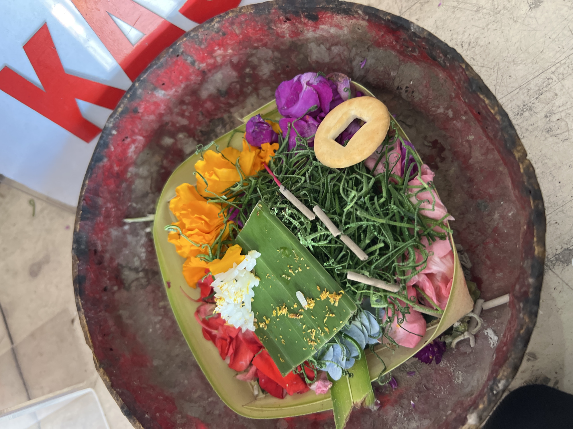 A Balinese offering, or Canang Sari, is displayed in a weathered, red-painted wooden bowl. The offering is placed on a woven palm leaf base and consists of various colorful flower petals, including red, pink, orange, purple, blue, and white. A small green leaf serves as a platform for some of the white petals and is sprinkled with what appears to be yellow zest. Two unlit incense sticks lie horizontally across the offering, and a light brown cracker-like object with a hole in the center rests on top of purple flower petals. Green, leafy material is abundant throughout the offering. The bowl itself shows signs of age and wear, with chipped red paint and a visible inner material. In the background, out of focus, is a white surface with large red letters, possibly part of a sign or banner, with the partial letter "K" visible. The setting appears to be outdoors, possibly on a textured concrete surface. The presence of the offering strongly suggests a location within Bali, Indonesia, likely in Denpasar where such religious practices are common.