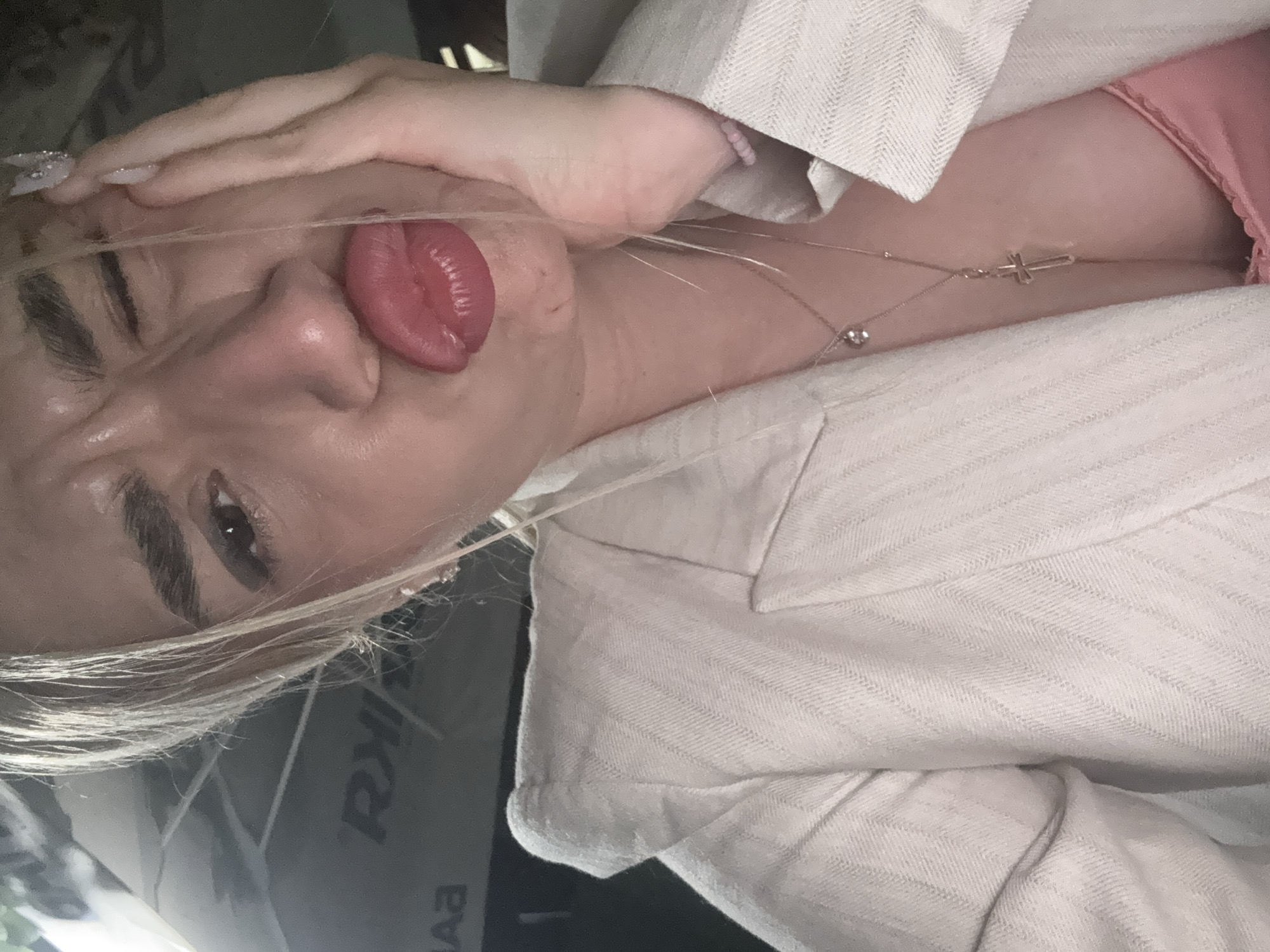 A medium-shot, vertically oriented selfie captures a young person from the chest up, with their head tilted to the right and eyes looking towards the camera. They have blonde hair that falls across their face, dark eyebrows, and their right eye is partially visible. Their left eye is winked shut. Their tongue is sticking out of their mouth. Their right hand is resting on the side of their head, near their temple, with long, light-colored nails visible. They are wearing a light-colored blazer or jacket with a herringbone pattern, and a pink top underneath, part of which is visible at the neckline. A delicate silver chain with a small cross pendant and another small, round charm hangs around their neck. The background is dark and appears to be indoors, possibly a room or vehicle, with some indistinct shapes and reflections. There is some faint, dark text visible in the background, appearing to be "RA" and "EV". The lighting is dim, suggesting it might be evening or indoors with artificial light. The person's expression is playful.