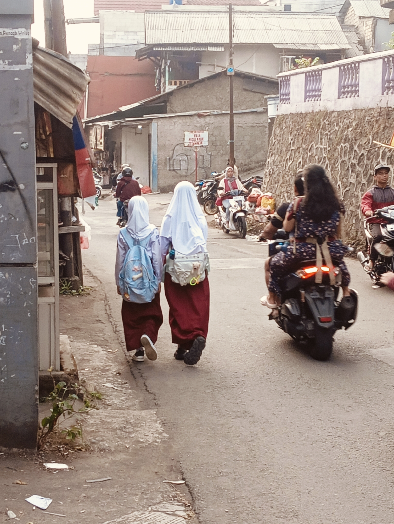 The image depicts a street scene in an urban environment. In the foreground, two young girls wearing school uniforms walk away from the camera, both with backpacks. The girl on the left has a light blue backpack, while the girl on the right has a white backpack adorned with a small stuffed animal. They are walking on a paved road with some debris and a small plant growing along the side.
Several people are on motorcycles in the mid-ground. A woman in a red and white outfit is riding a white motorcycle, and another person is riding a dark motorcycle behind her. Further back, a man in a red jacket is riding a motorcycle.
The background shows a mix of buildings, including houses with corrugated metal roofs and a stone wall with decorative purple railings. There is a sign visible on a building in the distance. The overall scene suggests a bustling, everyday street in a residential area. The location is identified as Unknown, Unknown.