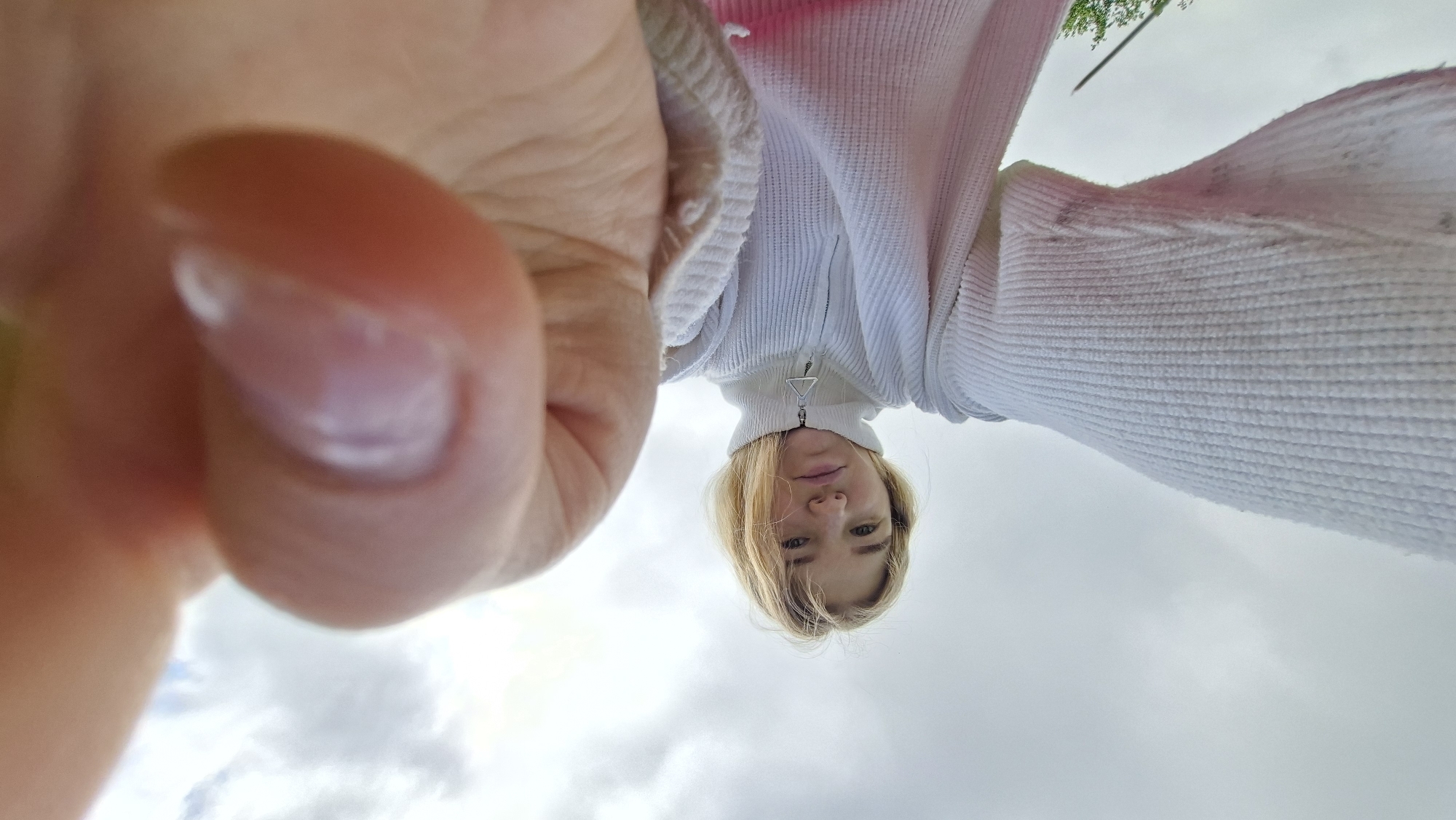 The image is a low-angle, close-up outdoor selfie featuring a young woman. The perspective is from below, looking upwards, causing the woman to appear inverted against the sky. Her right hand and thumb are prominently positioned in the left foreground, very close to the camera lens, showing clear detail of her fingernail.
The primary subject, a woman with blonde hair, is looking directly down towards the camera. She wears a light-colored, possibly white or off-white, ribbed long-sleeved top or jacket with a high collar and a central zipper. The zipper pull has a distinct metallic, somewhat triangular design. Her expression is neutral.
The background is dominated by a cloudy sky, suggesting an overcast or partly cloudy day. A small hint of green foliage, likely from a tree, is visible in the upper right corner, indicating an outdoor setting. There is no visible text in the image. The unique angle provides an intimate and unusual view, focusing heavily on the hand holding the camera and the subject looking down into it.