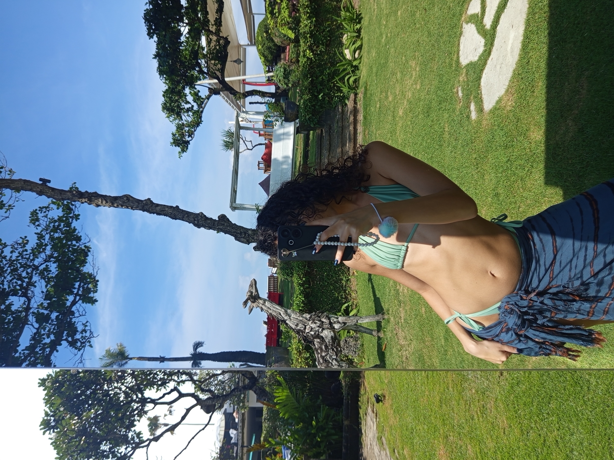 A woman is taking a selfie in front of a reflective panel or mirror. She is wearing a light green bikini top and a dark blue and brown wrap. She is holding a black smartphone with a pearl bracelet attached. The background shows a green lawn, trees, and what appears to be a building or structure with white canopies. A giraffe sculpture and what looks like a playground or recreational area are visible in the background. The reflection in the panel shows trees, a blue sky, and elements of the environment that are behind her. The location is identified as North Kuta, Indonesia.