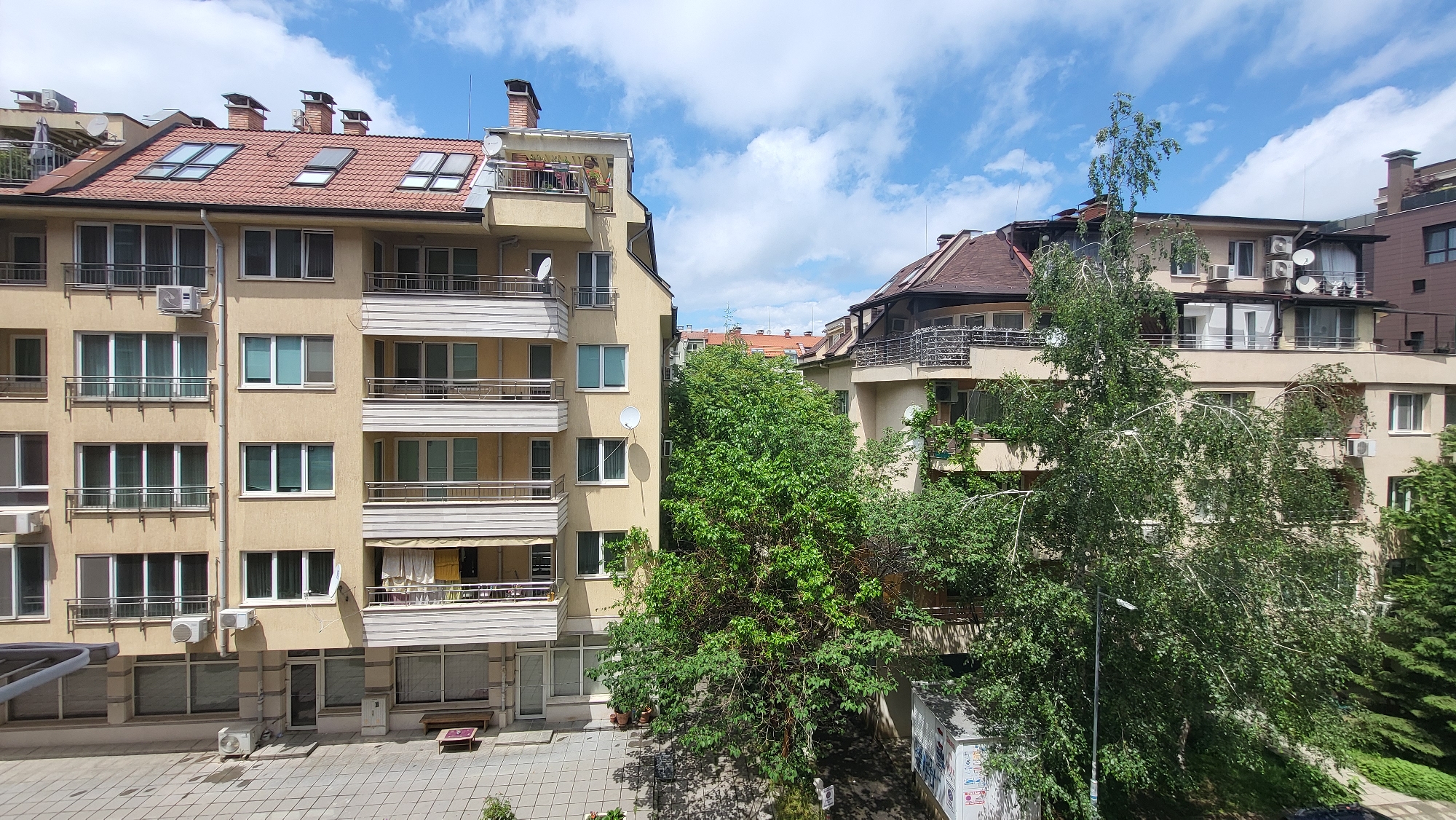 This image presents a bright, sunny daytime view of a residential area in Sofia, Bulgaria. The scene is dominated by multi-story apartment buildings and lush green trees under a blue sky with scattered white clouds.
On the left, a prominent light yellow/beige apartment building features numerous windows, many with railings, and multiple balconies, some open with metal balustrades and others enclosed. Several air conditioning units are affixed to its facade and balconies. The building is capped with a terracotta-tiled roof, punctuated by several skylights and chimneys. A satellite dish is also visible on one of its upper balconies. One balcony on a lower floor has a striped awning deployed, and laundry appears to be hanging to dry, suggesting typical residential activity.
In the center and extending to the right, a significant amount of vibrant green foliage from large trees partially obscures other buildings. A particularly tall tree with lighter branches and dense leaves stands on the right, while a darker green, bushier tree is positioned more centrally. Beyond the trees, another residential building, similar in style with multiple balconies, air conditioning units, and satellite dishes, and featuring a dark brown roof, is visible on the right side of the frame.
The ground area in the foreground at the bottom left shows a paved courtyard or sidewalk made of square tiles, where a small bench is present. A white kiosk or utility box, covered in what appears to be graffiti or posters, is partially visible on the right edge of this paved area.
No specific human figures are clearly visible, and no dynamic events are taking place, depicting a calm urban environment. The bright natural light, shadows, and clear sky indicate it is midday or early afternoon on a pleasant day. No discernible text is clearly readable in the image.
