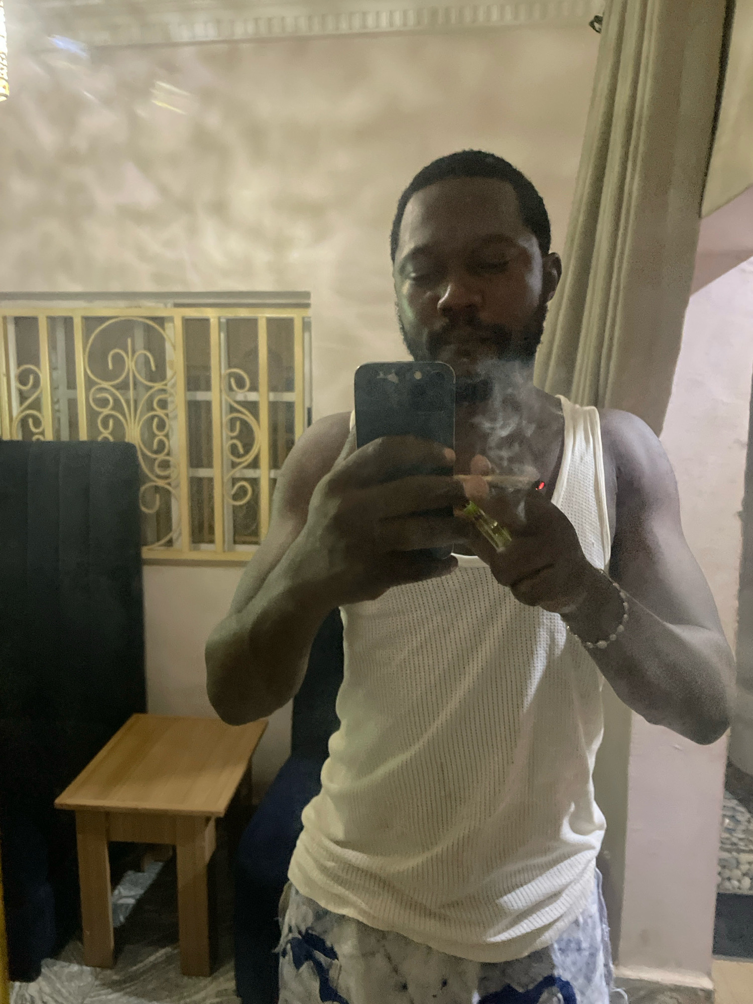 A man in a white tank top and patterned shorts is reflected in a mirror, holding a phone and a lit cigarette. He has a beard and is exhaling smoke. To his left, a black headboard is visible, and in front of it, a small wooden table. Behind the man, a window with decorative metal bars is present. The environment appears to be an indoor space, possibly a room in Abuja, Nigeria.