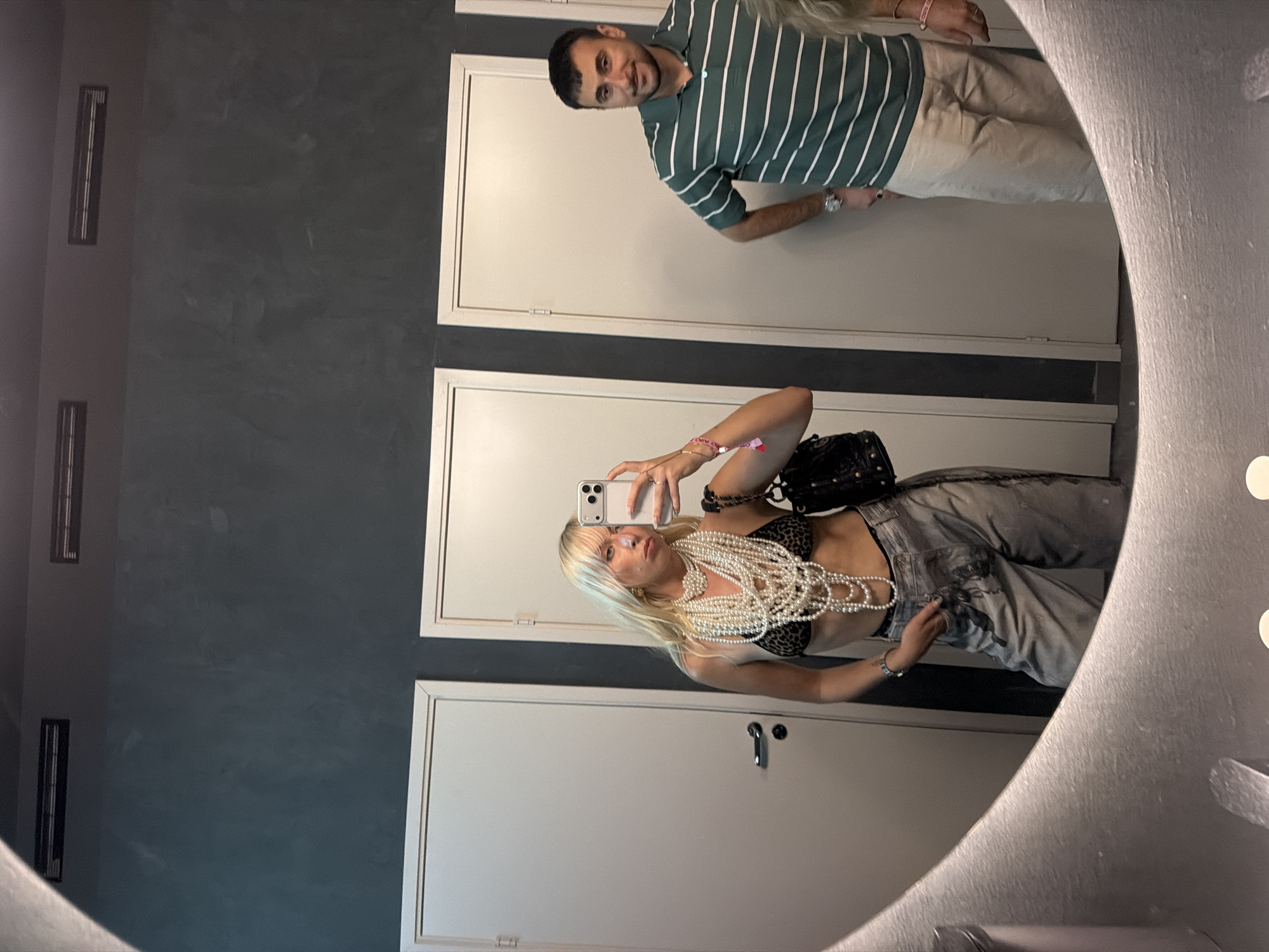 An indoor mirror selfie taken in an establishment in Huddinge, Sweden, depicts two individuals. A blonde female stands in the foreground, reflected in a large, irregularly shaped mirror that dominates the lower half of the frame. She wears multiple voluminous strands of white pearl necklaces over a leopard print bra top and light-colored, possibly distressed or paint-splattered, wide-leg trousers. She holds a light-colored smartphone in her right hand, capturing the reflection, and has her left hand in her pocket. A black studded handbag with a chain strap is visible on her left side, and a pink wristband and silver watch adorn her left wrist. Her facial expression is a slight pout.
Behind her, also reflected in the mirror, stands a male with short, dark hair, wearing a green polo shirt with horizontal white stripes and light-colored trousers. He stands with his right hand in his pocket, looking towards the camera with a slight smile.
The background consists of dark gray, textured walls and three white, plain doors with silver handles. Above the doors, two rectangular air vents are visible on the upper dark ceiling. The mirror's lower edge, visible in the foreground, shows two circular reflections of overhead lights and surface imperfections.