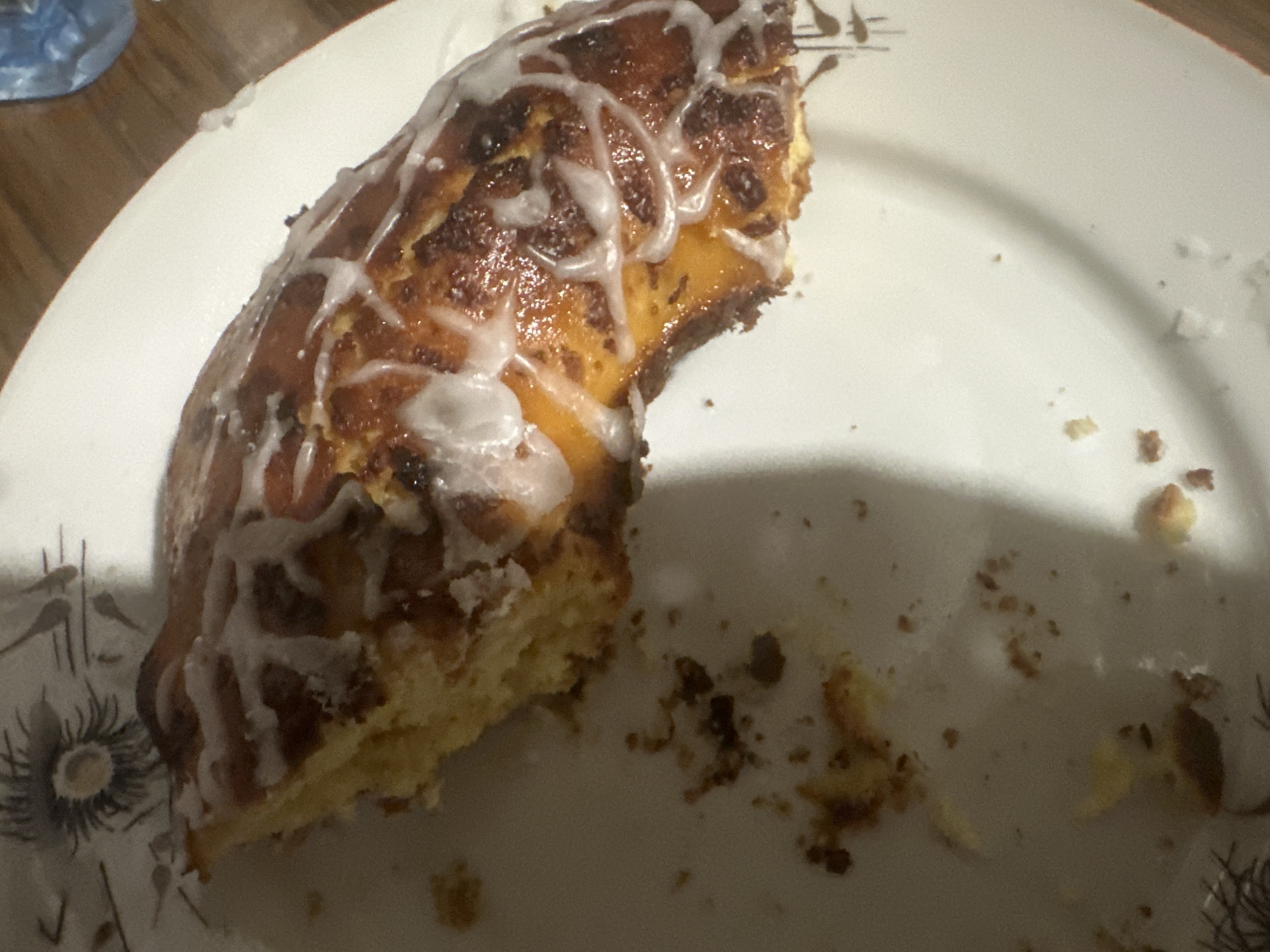 A partially eaten ring of pastry is on a white plate with decorative floral patterns. The pastry is drizzled with white icing and has a browned top. Crumbs are scattered around the remaining portion of pastry on the plate. The image does not depict or imply a location in Sirakovo, Bulgaria, or any other specific geographic location. There are no people depicted in the image.