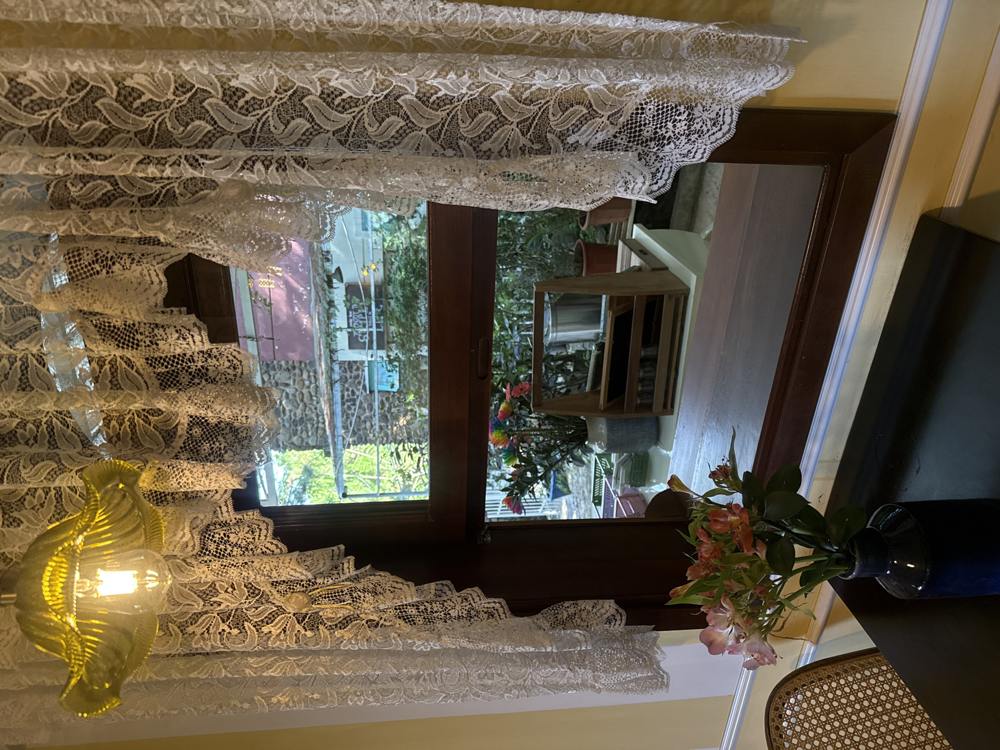This image depicts an indoor scene with a view through a window to an outdoor area. In the foreground, a dark tabletop holds a dark blue vase filled with a bouquet of pink and orange flowers. To the left of the table, a portion of a woven chair is visible. The window itself is framed by dark wood and adorned with white lace curtains. Through the window, an outdoor scene reveals greenery, a stone-clad building with a pinkish roof, and a blue sky. Inside the window, on a white surface, rests a wooden organizer with compartments, and a blue and white vase containing a colorful, rainbow-hued floral arrangement. Above the window, a yellow glass lampshade with an exposed lightbulb hangs. The overall impression is of a cozy, slightly vintage interior looking out onto a pleasant, possibly garden-like, outdoor setting. The specific location in Da Lat, Vietnam, is not directly discernible from the image itself, but the style and vegetation might suggest a residential or cafe setting typical of the region.