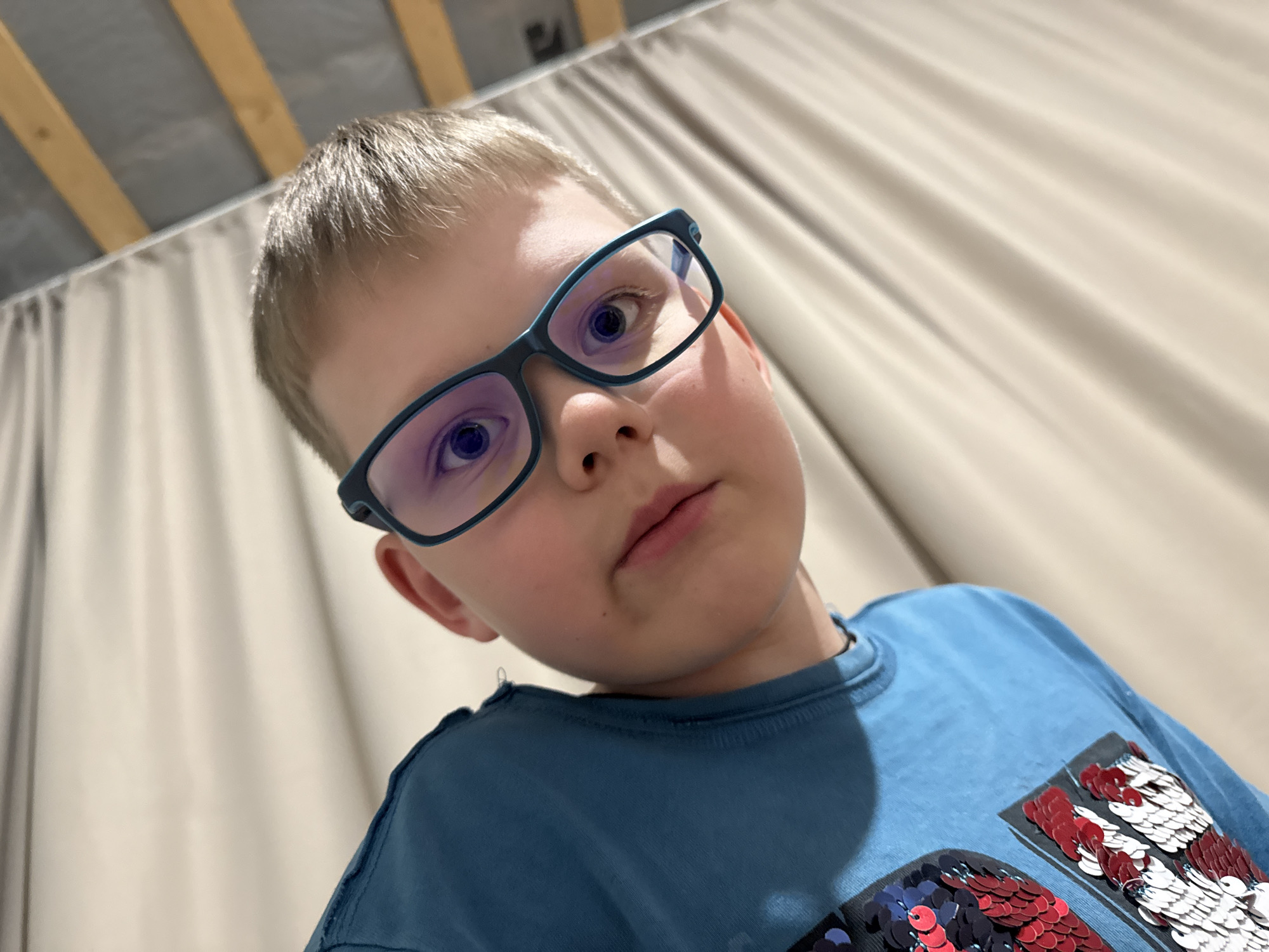 A young boy with short, light-colored hair and blue-framed glasses is featured in a close-up, low-angle portrait, looking directly at the camera. His eyes, magnified by the glasses, appear blue or violet, possibly due to reflections or lens tint. He wears a blue long-sleeved t-shirt with a distinctive sequined design on the front, composed of red, white, and dark blue/black sequins.
The setting is indoors, likely a room in Gornyi (Dvurechenskoe S/P), Russia. The background features light-colored fabric, resembling curtains or drapes, with visible vertical folds. Above this fabric, an exposed ceiling structure is visible, comprising light-colored panels or insulation alongside prominent wooden beams or rafters. The overall lighting appears to be artificial, illuminating the boy's face and upper body, which fill a significant portion of the frame. The boy's expression is neutral and direct. There is no visible text in the image.