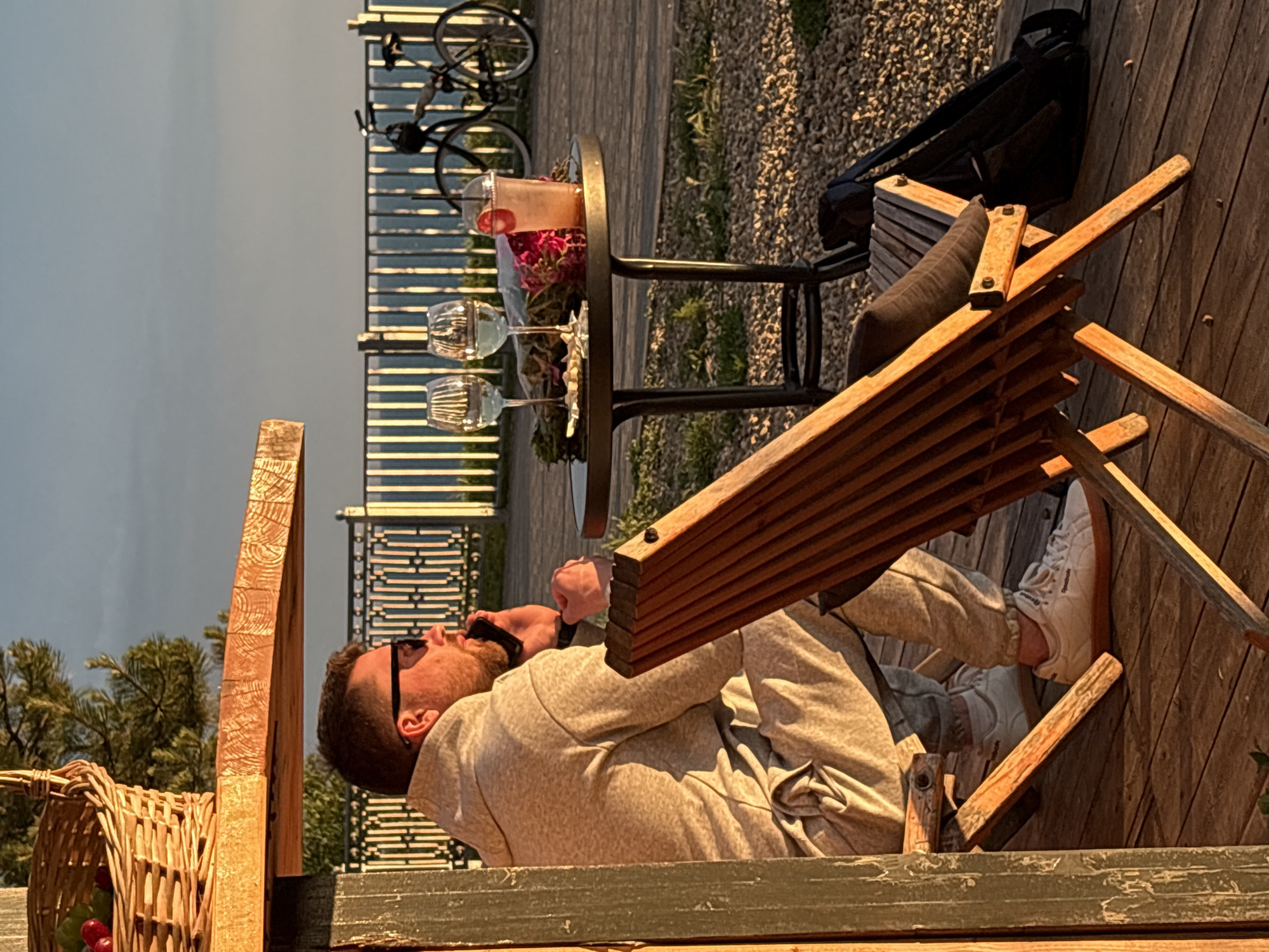 An outdoor scene depicts a man relaxing in a wooden slatted lounge chair on a wooden deck during the golden hour, likely late afternoon or early evening. The man, with a beard and wearing sunglasses, a light-colored long-sleeved top, and matching pants, holds a phone or small object to his ear. He wears white "Reebok" brand sneakers with gum soles.
Beside him is a small, round black metal table holding a clear pitcher of a red drink, possibly sangria, garnished with fruit and a straw. Two wine glasses, partially filled, and a small dish with snacks are also on the table. A black backpack is placed near the chair. In the foreground to the left, a wicker basket filled with green and red fruit is visible.
The setting overlooks a scenic view, suggested by a metal railing in the background. Beyond the railing, a calm body of water (or a hazy sky) stretches to the horizon, with some distant trees and foliage on the left. The ground beyond the deck features gravel and patches of greenery. A bicycle is parked further back, near the railing. The lighting casts warm, long shadows, contributing to a peaceful and relaxed atmosphere. The scene suggests leisure time at a location like a riverside or lakeside resort or private residence in Sennoi, Russia.
