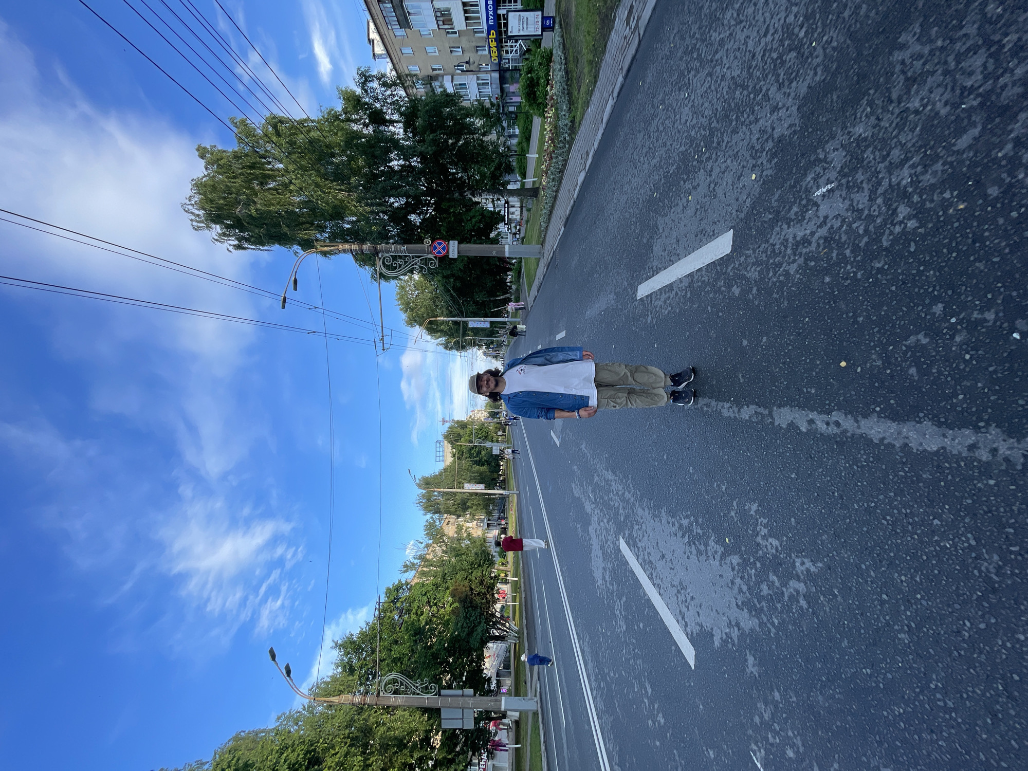 The image captures a street scene in a city, possibly after rain, as the asphalt appears wet and reflective. A man, wearing a light-colored t-shirt, a denim jacket, khaki pants, a baseball cap, and dark sneakers, stands in the middle of the road, facing the camera. He is the central subject. The road is wide, with multiple lanes marked by white lines. On either side of the road are sidewalks, trees, streetlights, and buildings. There are other people visible in the background, some walking on the sidewalks and one child in the distance. Overhead, there are power lines and a bright blue sky with scattered white clouds. The environment appears to be a typical urban street with residential and commercial buildings.