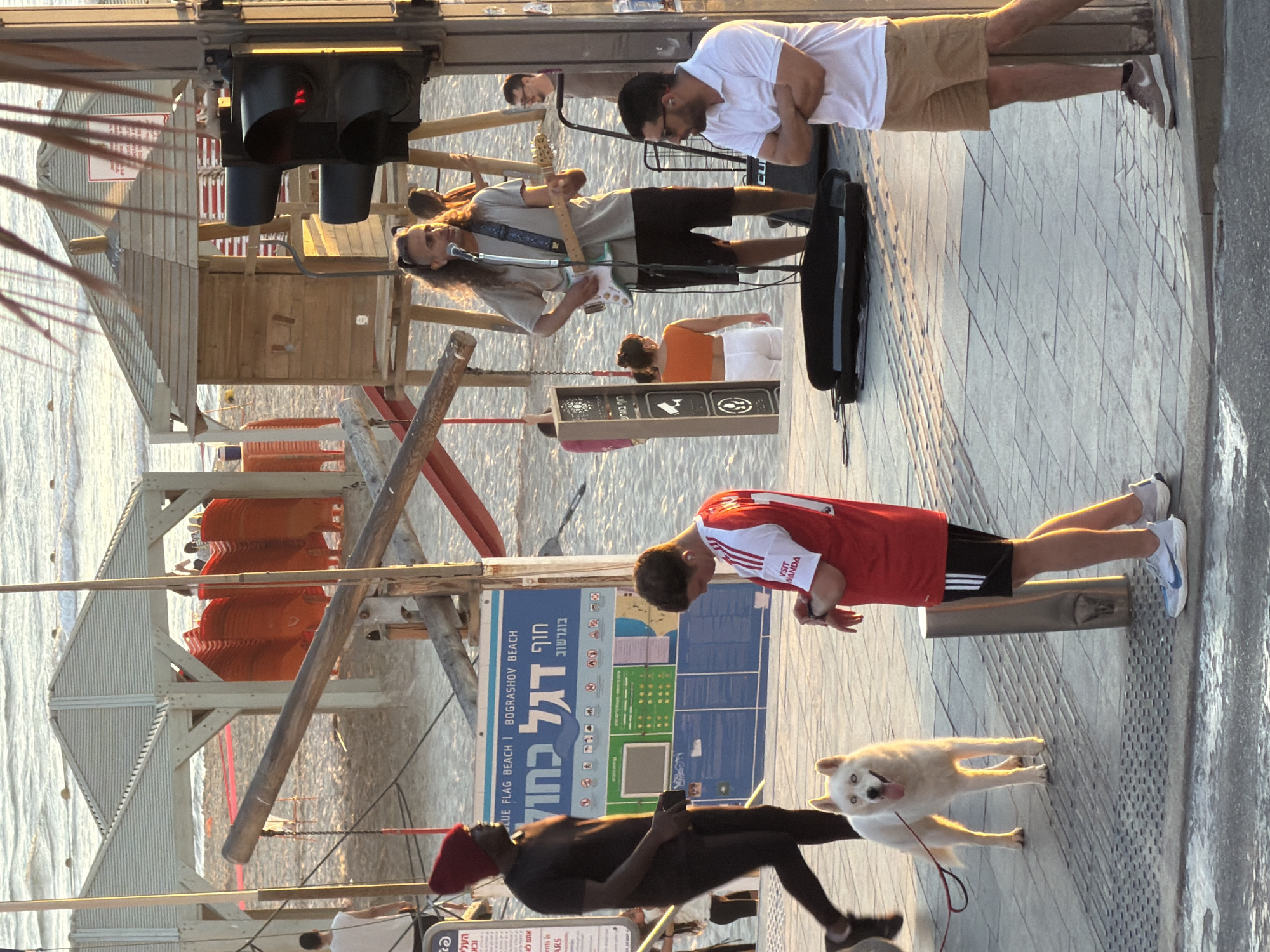 The image captures a vibrant scene at Bograshov Beach in Tel Aviv, Israel. A sandy beach stretches towards the sea, with several rows of orange beach chairs stacked on a platform. Along the paved walkway, a musician with long hair plays an electric guitar, accompanied by a man standing beside him. A person in a red shirt and black shorts stands by the beach information sign. A person walks on the pavement with a white dog on a leash. The beach scene is a mix of leisure, music, and urban life.