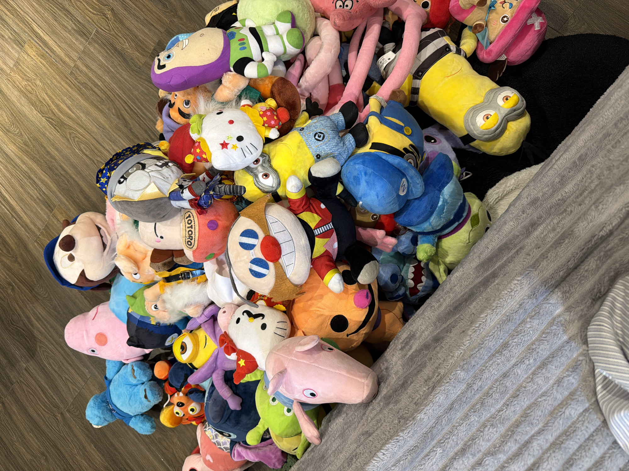 The scene depicts a large pile of assorted plush toys on a floor with light brown wood-grain laminate planks on one side and a gray, textured blanket on the other. The plush toys are a mix of licensed characters and generic designs, including Minions, Hello Kitty, Peppa Pig, Stitch, and characters from Pixar films. The plush toys appear to be densely packed.
There is no evidence suggesting this image was taken in Đà Nẵng, Vietnam.