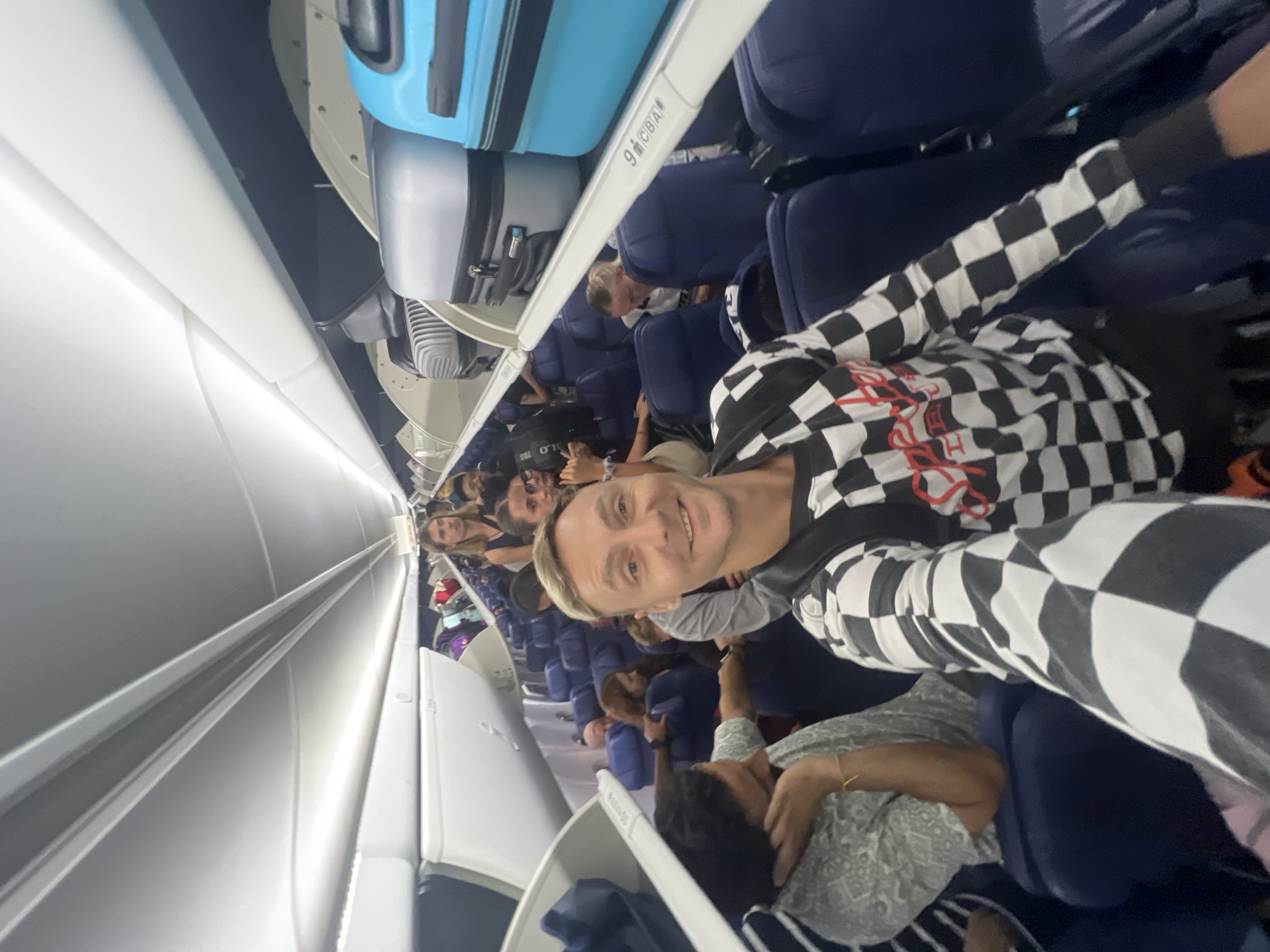 The media file presents a sideways-oriented view inside a commercial airplane cabin, likely associated with travel to or from Kuta, Indonesia. The main subject is a man in the foreground, appearing to take a selfie. He has light-colored hair and is smiling broadly at the camera. He wears a distinctive long-sleeved shirt with a black and white checkerboard pattern, featuring some red text on the chest.
Behind the man, numerous other passengers are visible seated in the blue airline seats lining the aisle. Their faces and expressions are less distinct, but they appear relaxed, some looking forward while others glance towards the camera. Above the passengers, the overhead bins are closed, with several pieces of luggage stored within, including prominent blue and grey suitcases. The white walls and ceiling of the aircraft, along with the cabin lighting, are also visible. On one of the overhead bins, the seating markers "9 C B A" are discernible. The scene captures a typical moment inside a passenger flight, conveying a generally calm and cheerful atmosphere, particularly from the smiling subject.