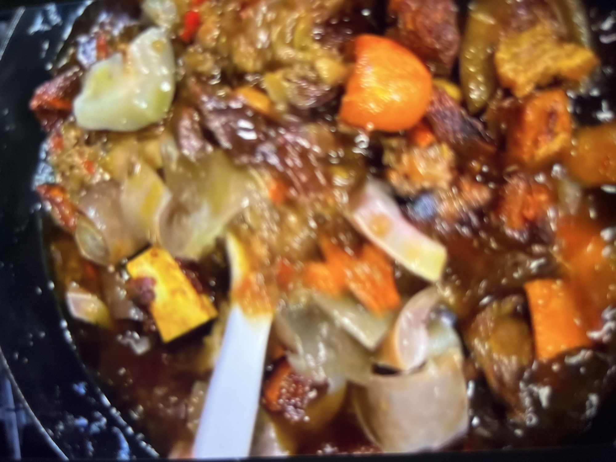 The image is a close-up, blurred shot of a pot filled with a dark, soupy stew. The stew contains various chopped ingredients, including what appear to be chunks of meat, root vegetables like carrots and possibly sweet potatoes (indicated by orange and yellow colors), and some lighter-colored, possibly starchy pieces. There are also small red pieces, which could be chilies or other spices. A white utensil, likely a spoon or spatula, is visible on the left side of the frame, partially submerged in the stew. The background is dark, suggesting the pot is on a stovetop or similar surface. The overall impression is of a hearty, home-cooked meal. There is no visible context to suggest a specific location in Abuja, Nigeria.