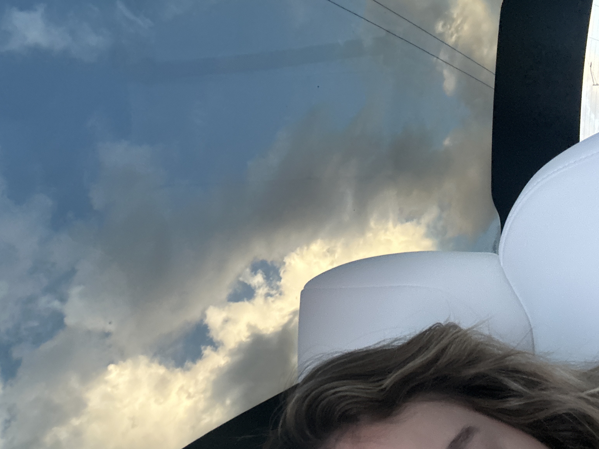 The image shows a partial view of a person's face and hair, with a white car seat in the foreground. The background is a cloudy sky, reflecting in the car's windshield or sunroof. The scene appears to be from inside a vehicle. The location is identified as San Bernardino Tlaxcalancingo, Mexico.