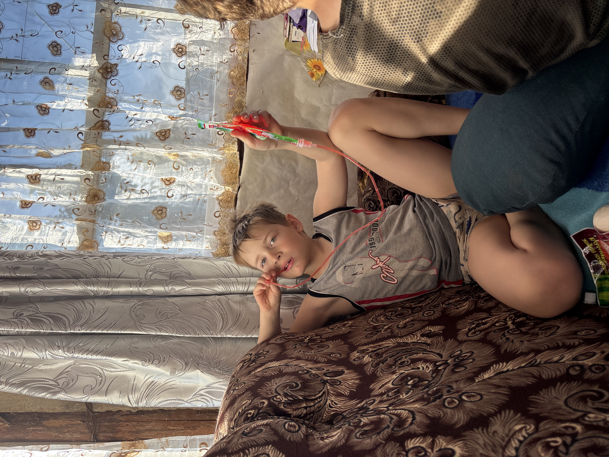 A young boy with blond hair and blue eyes lies on a brown patterned couch, holding a colorful toy connected to a red tube. He is wearing a grey t-shirt with red and black trim and patterned shorts. The boy looks towards the camera with a neutral expression. Sunlight streams through a sheer curtain with floral embroidery behind him, suggesting it's daytime. The room appears to be a living space, possibly in a home, with the couch as the central element. Another person's arm and shoulder are visible at the top of the frame, partially obscured. There is a glimpse of a blue blanket and a product package with Cyrillic text near the boy's legs. The overall scene is casual and domestic.
