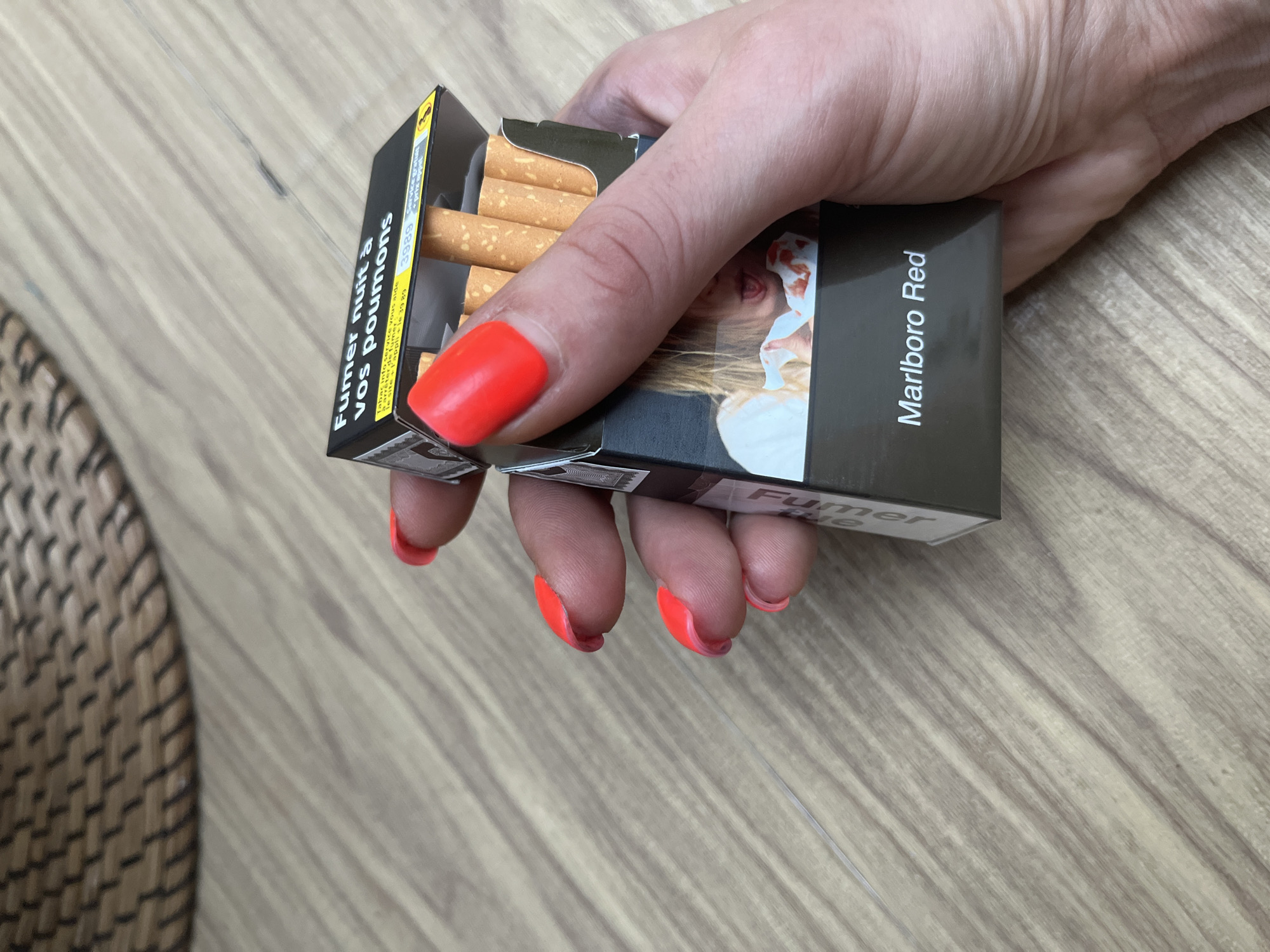A hand with bright red nail polish holds an open pack of Marlboro Red cigarettes. The pack displays a health warning in French, "Fumer nuit à vos poumons" (Smoking damages your lungs), and a graphic image of a child wiping blood from their nose. The background is a light-colored wooden surface, and the upper left corner shows a woven basket.