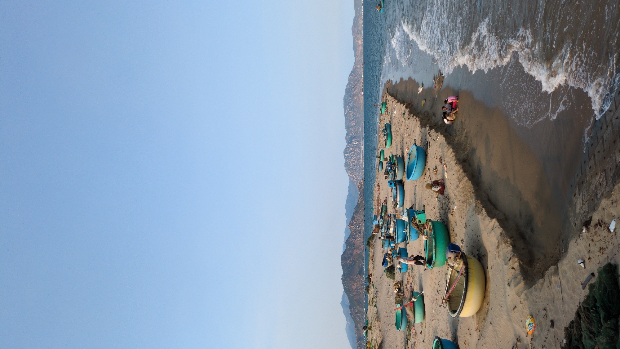 The image shows a coastal scene in Phan Rang–Tháp Chàm, Vietnam. Several round, colorful fishing boats are scattered along a sandy beach. People are visible near the shoreline. The beach transitions into the ocean, with waves coming ashore. Mountains or hills form a backdrop to the beach. The sky above is clear and blue.