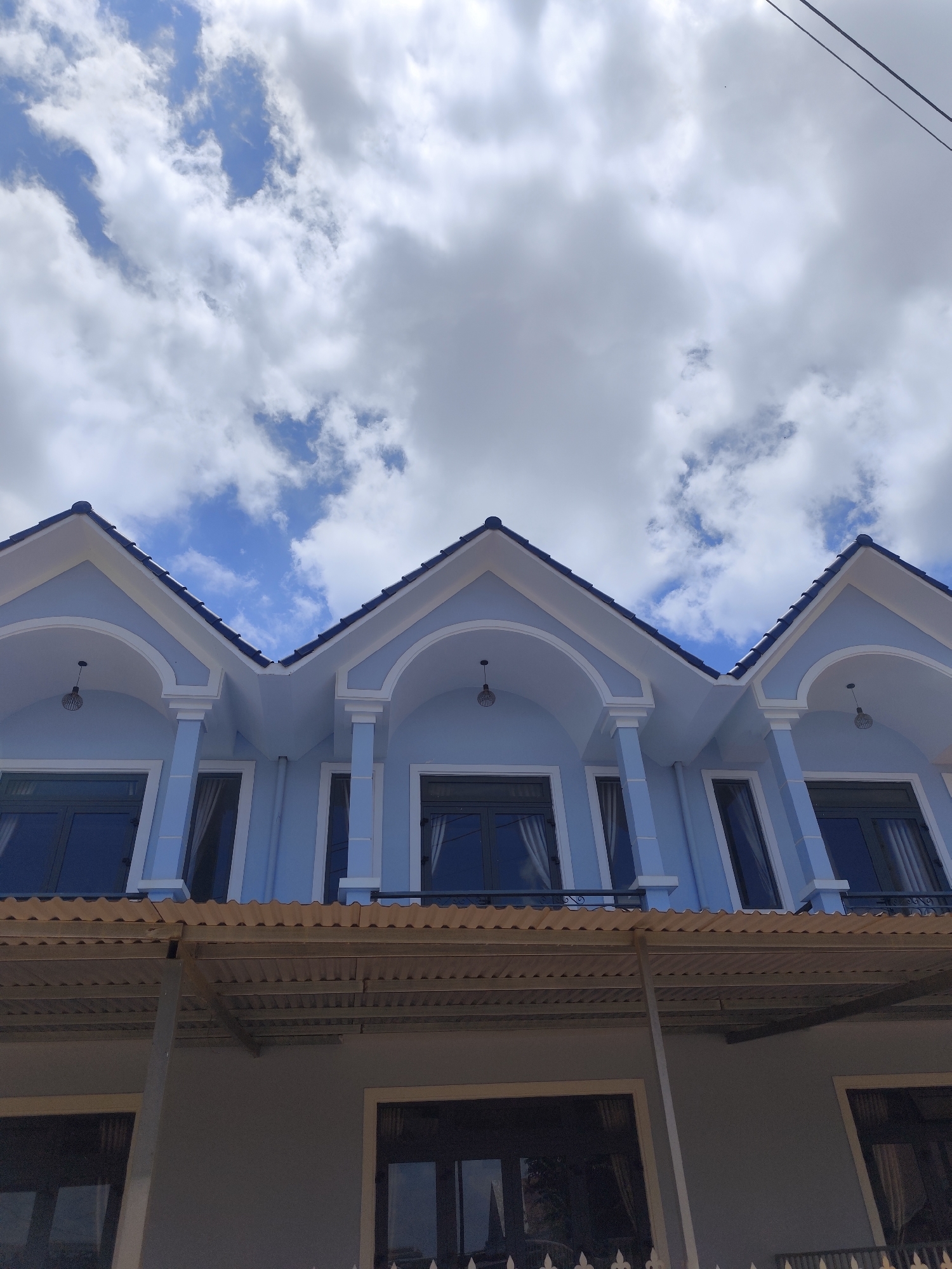 A low-angle, eye-level shot captures a bright light blue, two-story building under a cloudy sky. The architecture of the building's second floor is prominent, featuring three distinct sections, each topped with a dark blue tiled gable roof. White trim outlines these gables, and each section includes an arched facade in white, supported by white columns, creating recessed areas for windows. Dark-framed windows are visible within these arches, with some showing white curtains inside. Below the second story, a corrugated metal roof, supported by metal framing, extends outwards, likely serving as a cover for a lower balcony or ground floor area. A portion of the ground floor is visible, showing more dark-framed windows. The sky overhead is a bright blue, filled with numerous white and grey fluffy clouds. Thin, dark utility wires diagonally cross the upper right corner of the frame. The image depicts a modern-style building during the daytime under fair weather conditions in Lạc Dương, Vietnam. No people or visible text are present.