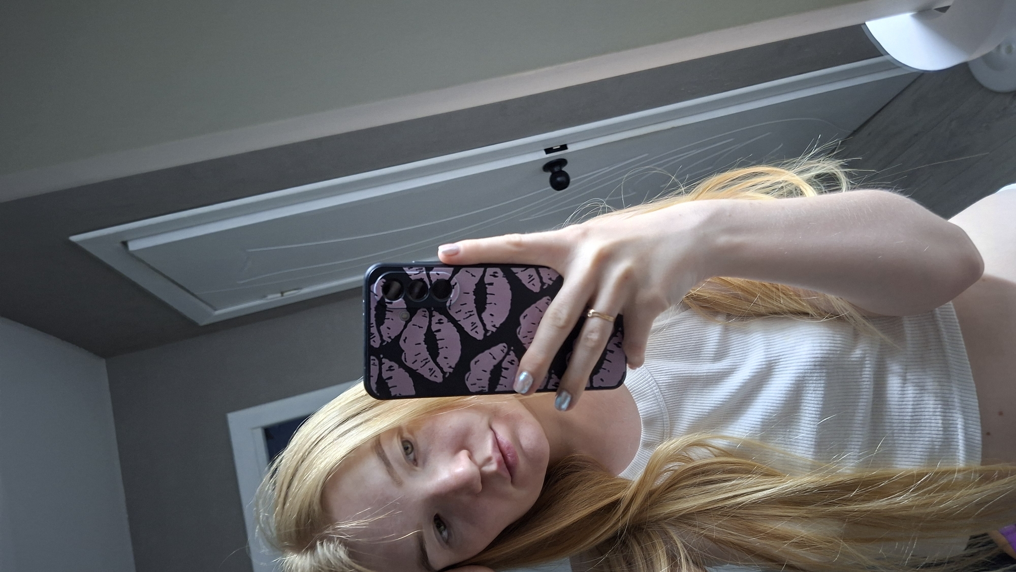 A young woman with long blonde hair is taking a selfie in what appears to be an indoor setting. She's holding a smartphone with a black case decorated with pink lip prints. She is wearing a white ribbed shirt. The background includes a white ceiling access panel. The location is unspecified; no information supports the suggestion that the image was taken in the city of Chaikovsky, Russia.