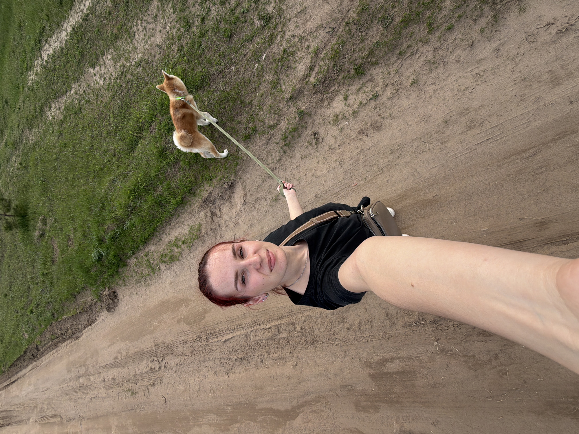 A selfie taken in Chaikovsky, Russia, shows a woman on a dirt road holding a leash attached to a dog. The dog, a tan and white Shiba Inu-type breed, is positioned to the woman's left, slightly off the road on a grassy area. The woman wears a black t-shirt and has a cross-body bag. The ground is mostly dirt with tire tracks, and some grass can be seen along the edges.