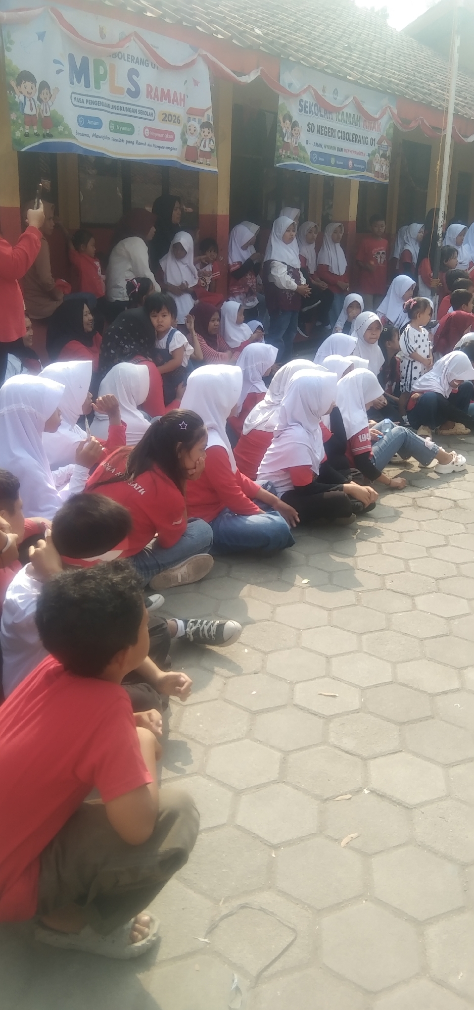In Rahayu, Indonesia, a group of children and adults are gathered outdoors, possibly for a school event. The scene is set in front of a building with a banner that reads "MPLS RAMAH" and "SEKOLAH RAMAH ANAK SD NEGERI CIBOLERANG 01". Many of the children are wearing white or red clothing and headscarves, suggesting a school uniform or participation in a specific event. Some are sitting on the ground, while others are standing or watching. The ground is paved with hexagonal tiles. The overall atmosphere appears to be one of a community gathering or educational activity.
