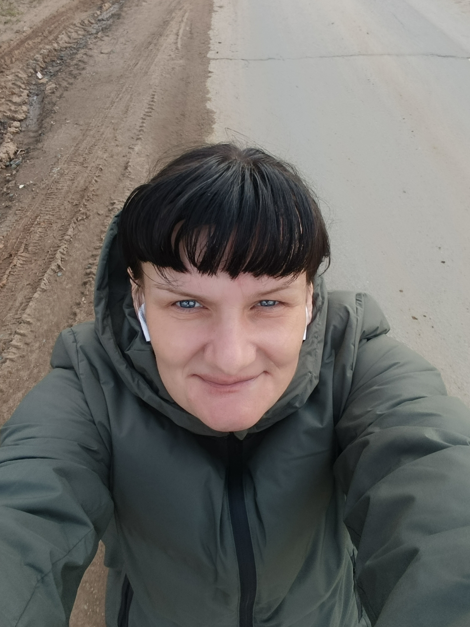 A woman takes a high-angle selfie outdoors in Фоки, Russia. She has dark hair styled with bangs and prominent blue eyes. She is wearing a dark green hooded puffer jacket, zipped up, and has white wireless earbuds in both ears. Her expression is pleasant and calm, featuring a subtle smile as she looks directly into the camera.
The background behind her shows a distinct road with two different surfaces. To her left (viewer's right), the ground is an unpaved, muddy track, deeply rutted with tire marks and some scattered debris. To her right (viewer's left), the road transitions to a lighter-colored paved or concrete surface, which appears mostly flat but shows some minor cracks and discoloration. The lighting suggests it is daytime, likely under overcast conditions, as there are no strong shadows. The overall scene captures a casual moment in a rural or semi-rural outdoor setting. No visible text is present in the image.