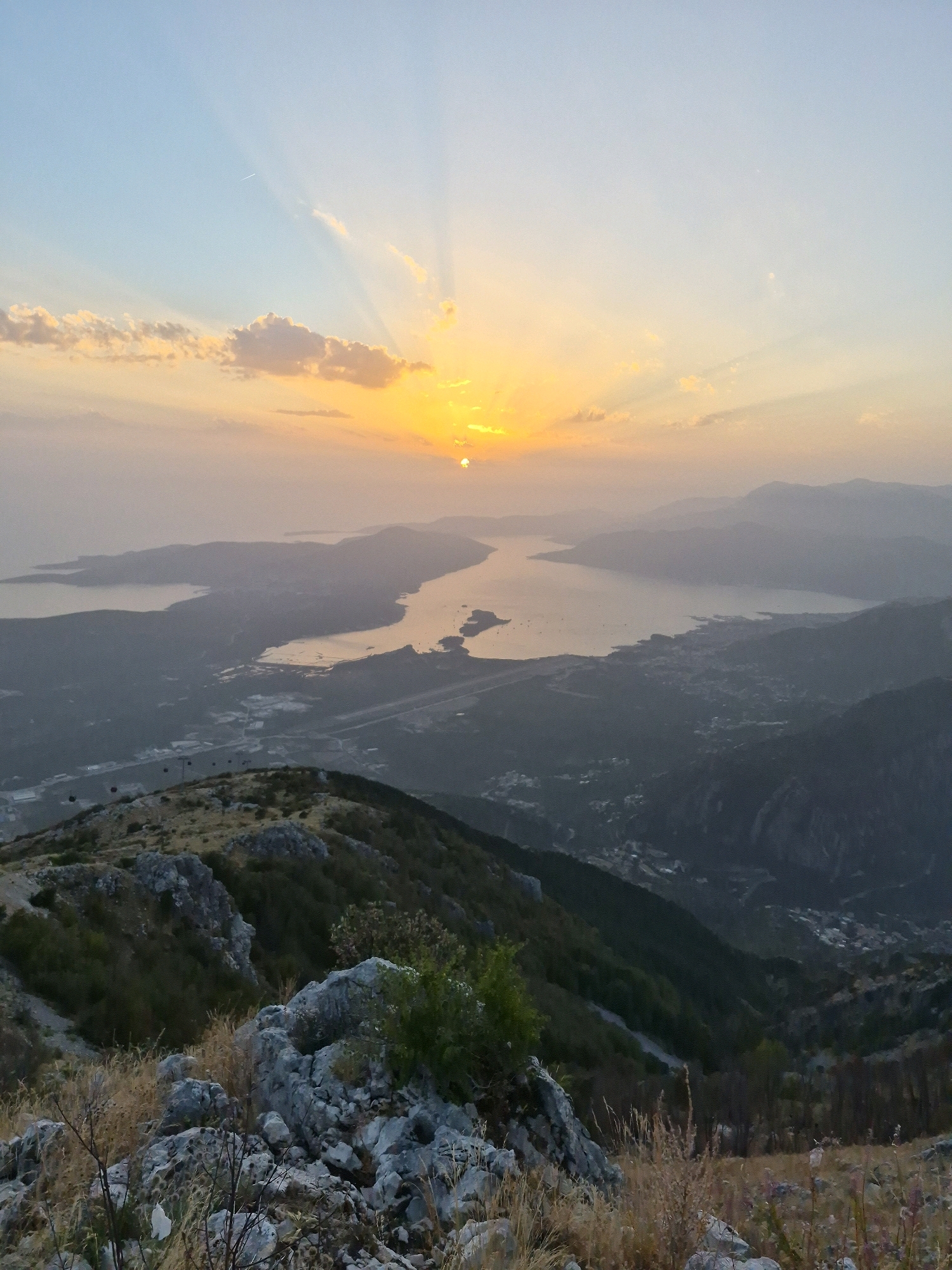 This image captures a panoramic view of the Bay of Kotor in Montenegro, as seen from a high vantage point, likely near the city of Kuk. The foreground is dominated by a rocky, grassy hillside with dry vegetation and scattered boulders. In the midground, the expansive bay stretches out, its waters reflecting the warm hues of the setting sun. The distant landscape is characterized by rolling hills and mountains, shrouded in a light haze. A cable car line with several cabins is visible traversing the hillside in the midground, suggesting a tourist attraction. The sky above is a gradient of soft blues and oranges, with the sun partially obscured by thin clouds, casting ethereal rays across the scene. The overall impression is one of serene natural beauty, with a hint of human presence through the cable car infrastructure.