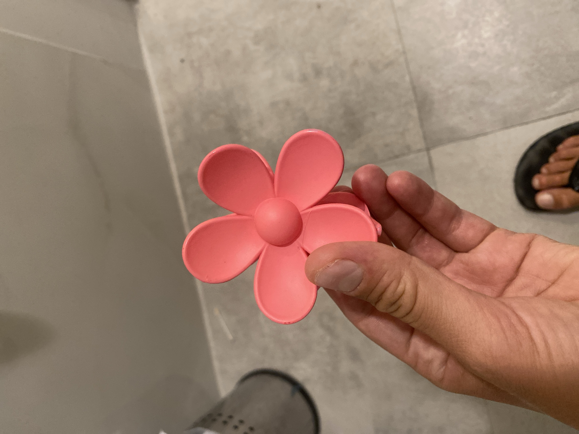 A close-up, eye-level, full shot shows a person's hand holding a pink flower-shaped hair clip. The hair clip has five petals and a round center. The background consists of a tiled floor and a wall. The setting is a bathroom. The image was taken in Aksu, Turkey.