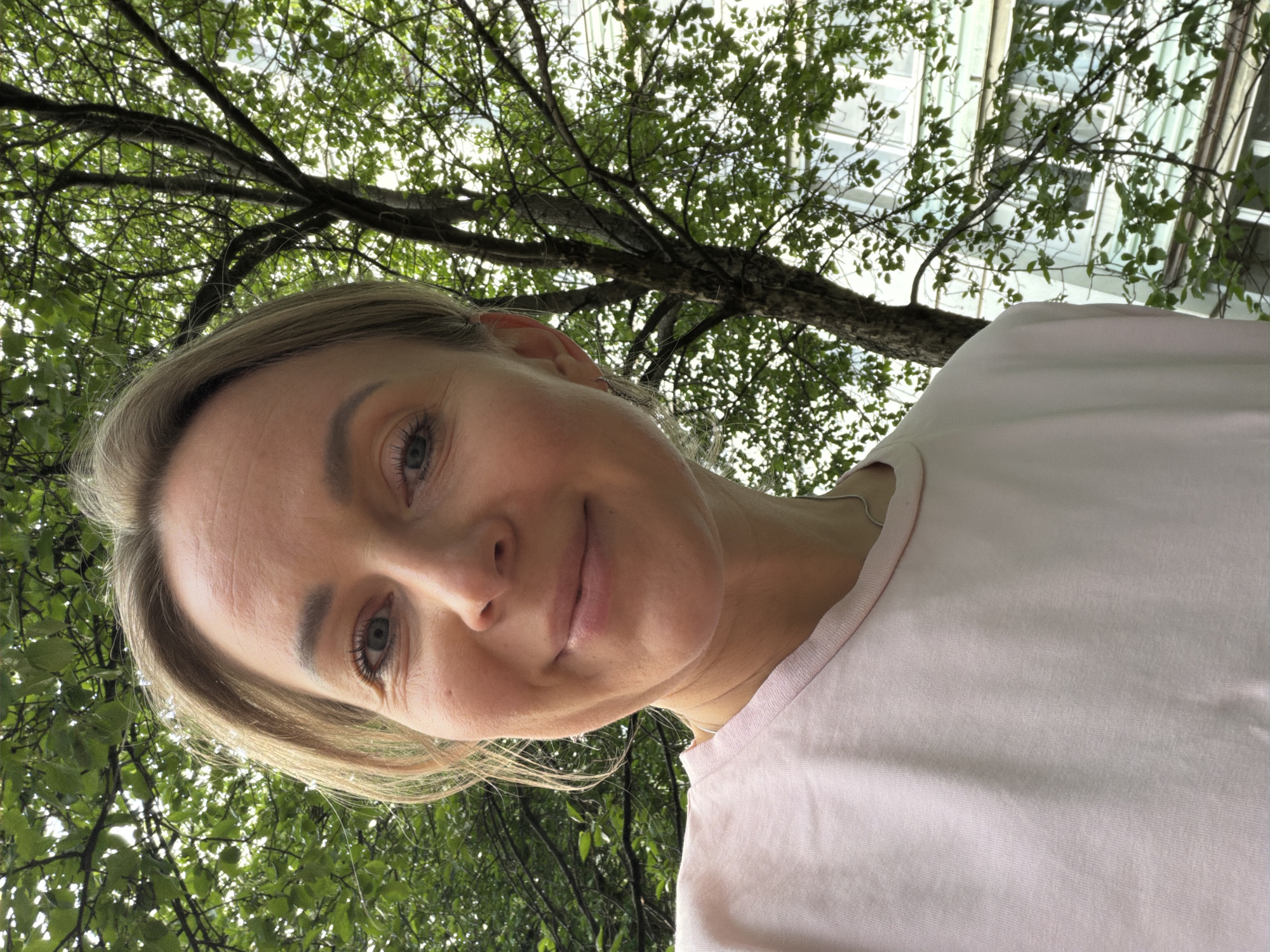 A woman with light skin, blonde hair, and blue eyes is pictured from a low angle. She is wearing a light pink shirt. Behind her is a tree with green leaves and branches. A building is visible through the branches of the tree. The location is likely Chaikovsky, Russia. The image is a selfie taken outdoors.