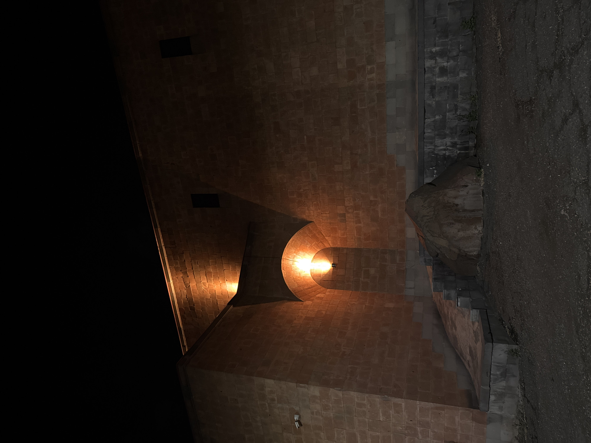 The image shows a night scene in Yerevan, Armenia, featuring a brick structure illuminated by warm lights. The structure appears to be part of a building or an archway, with its bricks creating a textured surface. On the right side, there is a rough, dark stone or concrete wall and a street-level surface. The left side of the image is mostly dark, suggesting the structure extends into the night sky or an unlit area. The overall impression is that of an urban environment, with architectural details highlighted by artificial lighting.