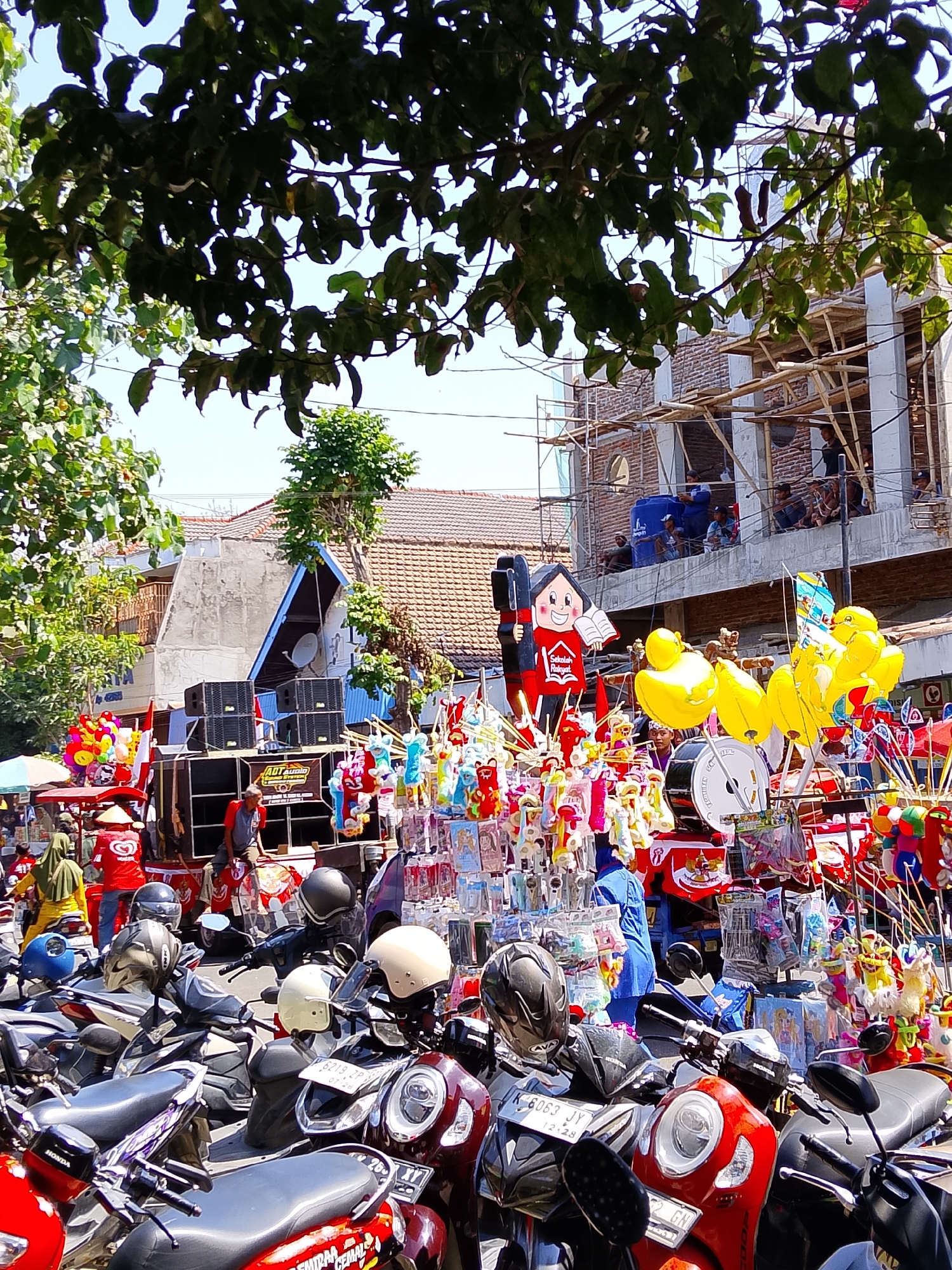 The image captures a lively outdoor market scene in the city of Ngelo, Indonesia. In the foreground, a cluster of parked motorcycles suggests a busy area where people have arrived to shop or attend an event. The motorcycles are of various colors and models, with some adorned with helmets.
Behind the motorcycles, a vibrant display of market stalls is visible. These stalls are laden with an array of colorful goods, including balloons, stuffed toys, and various other trinkets. A prominent stand features a large cutout figure of a child reading a book, with the text "Sekolah Rakyat" (People's School) on its chest, indicating it might be related to an educational or community event.
Further into the background, a large sound system with speakers is set up, implying that music or announcements are part of the event. A man in a red shirt is seated near the sound system, possibly involved in its operation. Adjacent to this, a building is under construction, with scaffolding and brickwork visible, suggesting ongoing development in the area. People are seen on different levels of the construction site, observing the market below.
The scene is illuminated by bright daylight, with shadows cast by trees and structures, indicating an outdoor setting. The overall atmosphere appears to be one of community gathering and festive activity, possibly a local festival or market day. The presence of diverse elements like the market stalls, sound system, and construction site paints a picture of daily life and development within Ngelo.
