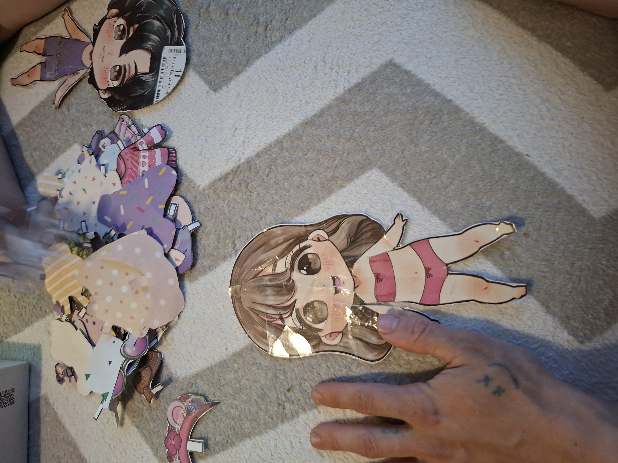 The image shows a collection of anime-style paper dolls and clothes laid out on a gray and white chevron patterned surface. A person's hand is near one of the dolls. A box is visible in the lower left corner of the image. The location of the image is Unknown, Unknown.