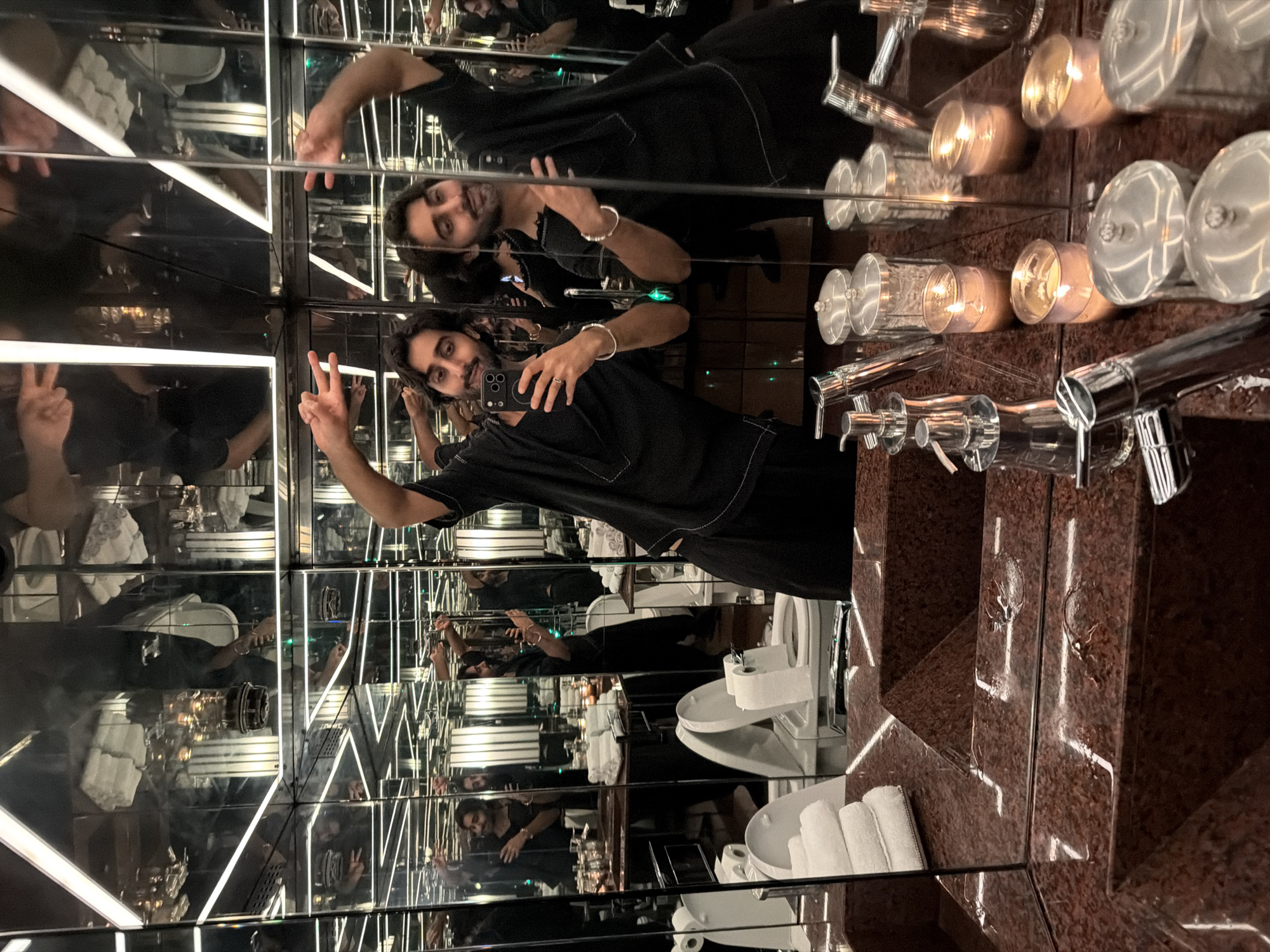 The image shows a man taking a selfie in a room with mirrored walls and ceiling, creating a disorienting, infinity-room effect. The man is wearing a black outfit and is holding a smartphone in his right hand. He is making a peace sign with his left hand. Multiple reflections of the man and the room are visible due to the mirrored surfaces.
The room appears to be a bathroom or a dressing area, with a toilet, sinks with faucets, and neatly folded white towels visible in the reflections. There are also several glass candle holders with lit candles, casting a warm glow. The walls of the vanity area are made of a dark, speckled material, possibly granite. The overall lighting is dim, with the main light sources being the candles and the bright white strip lights integrated into the ceiling and walls. The reflections suggest the presence of other people in the room, though they are not clearly visible. The man's expression appears relaxed. The scene is indoors, and there are no obvious cues about the time of day or weather outside. The location is identified as London, United Kingdom, but this context doesn't add specific visual details to the description of the image itself. There is no visible text in the image.