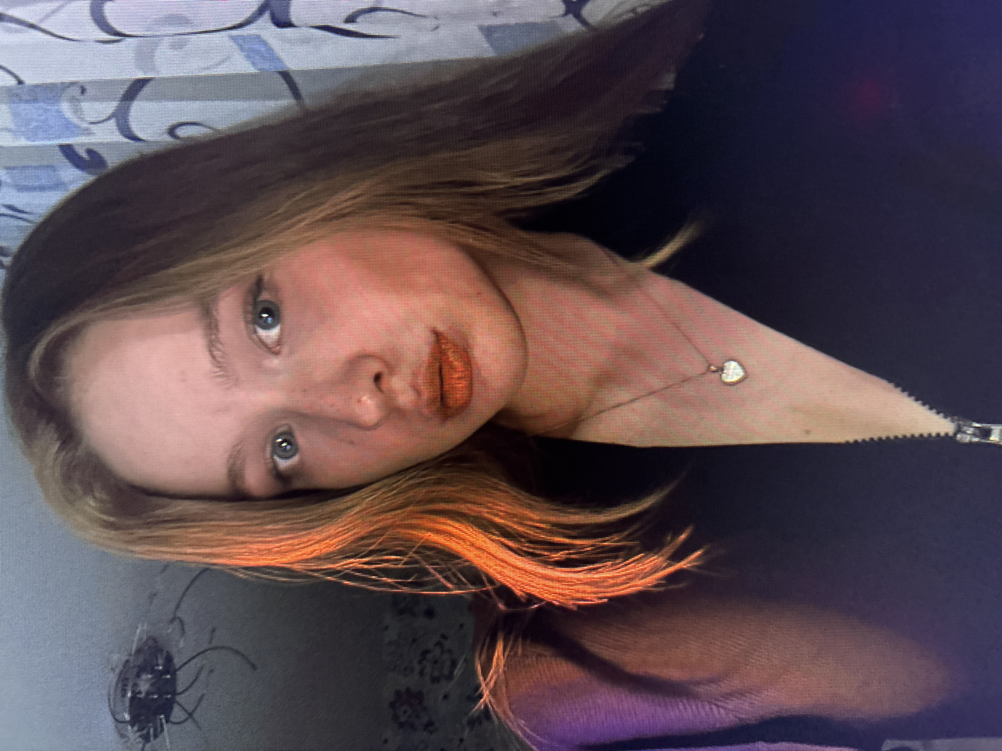 A rotated close-up image shows a young woman with light skin, blue eyes, and medium-length brown hair with golden highlights at the ends. She is wearing a black top with a visible zipper pull and a delicate necklace with a heart pendant. Her makeup includes a nude lip color. The background includes a wall with a pattern of white and blue, and a section of a table with a dark floral or patterned covering that also reveals some reflection. No location is specified, but the user suggested Chaikovsky, Russia.
