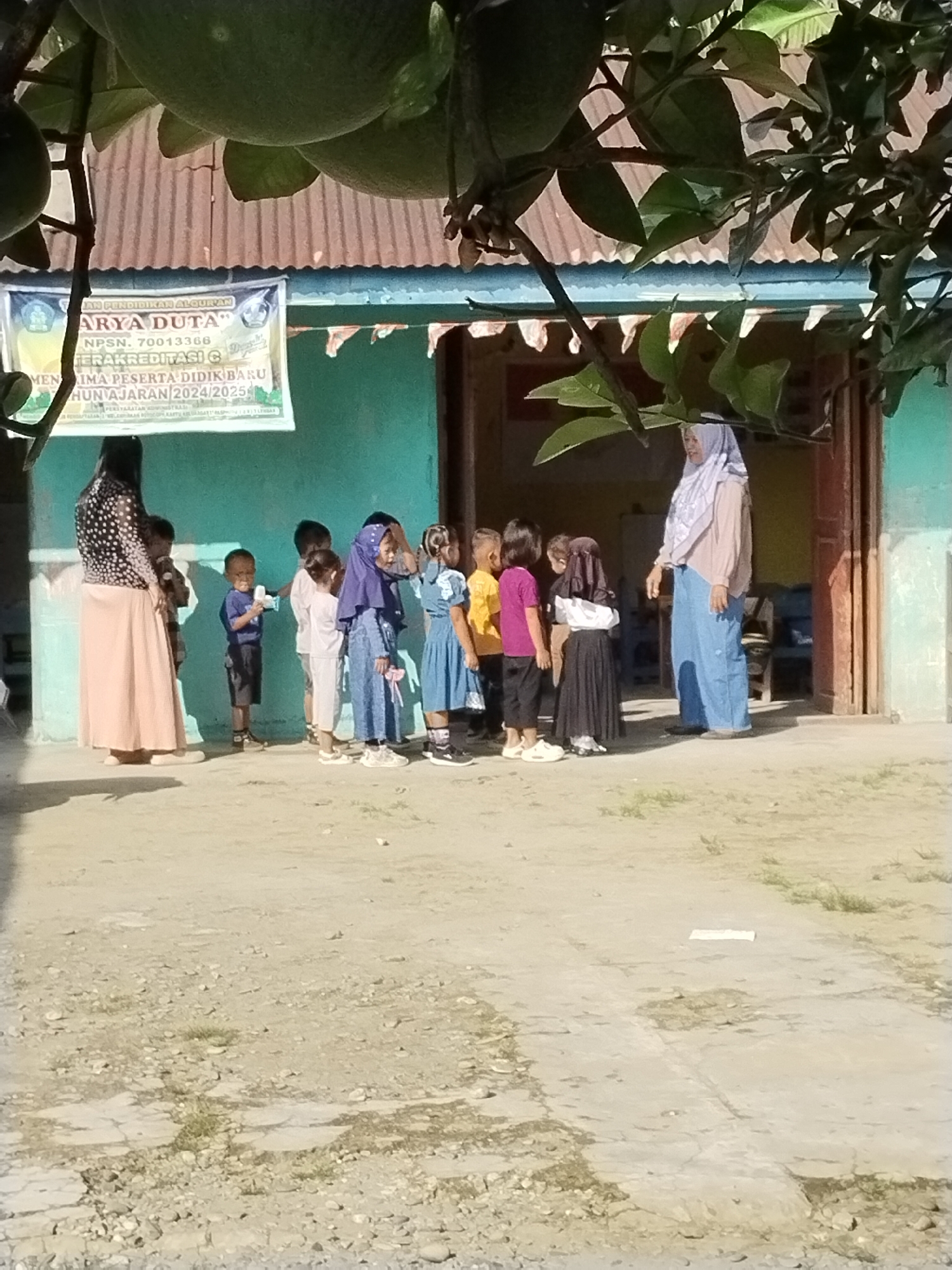 In the city of Sawit Seberang, Indonesia, an outdoor scene unfolds near a turquoise-colored building. The foreground is dominated by dry, sandy ground with scattered small stones and patches of dry grass. Overhead, large green fruits hang from branches, partially obscuring the view.
A group of children, predominantly young, are gathered on the ground. Several are wearing uniforms: some in blue dresses, one in a purple headscarf and long skirt, and others in casual shirts and pants. A woman in a light brown headscarf and blue pants stands near the entrance of the building, appearing to address the children. Another woman, wearing a black and white polka dot top and a long pink skirt, stands to the left, facing away from the camera. A banner is visible on the building's exterior, displaying text in Indonesian, indicating it is an educational institution, specifically "PAUD" (Pendidikan Anak Usia Dini, early childhood education) and mentioning "KARYA DUTA" and school year information. The building itself has a corrugated metal roof and wooden doorframes. The overall atmosphere suggests a school environment during the daytime.