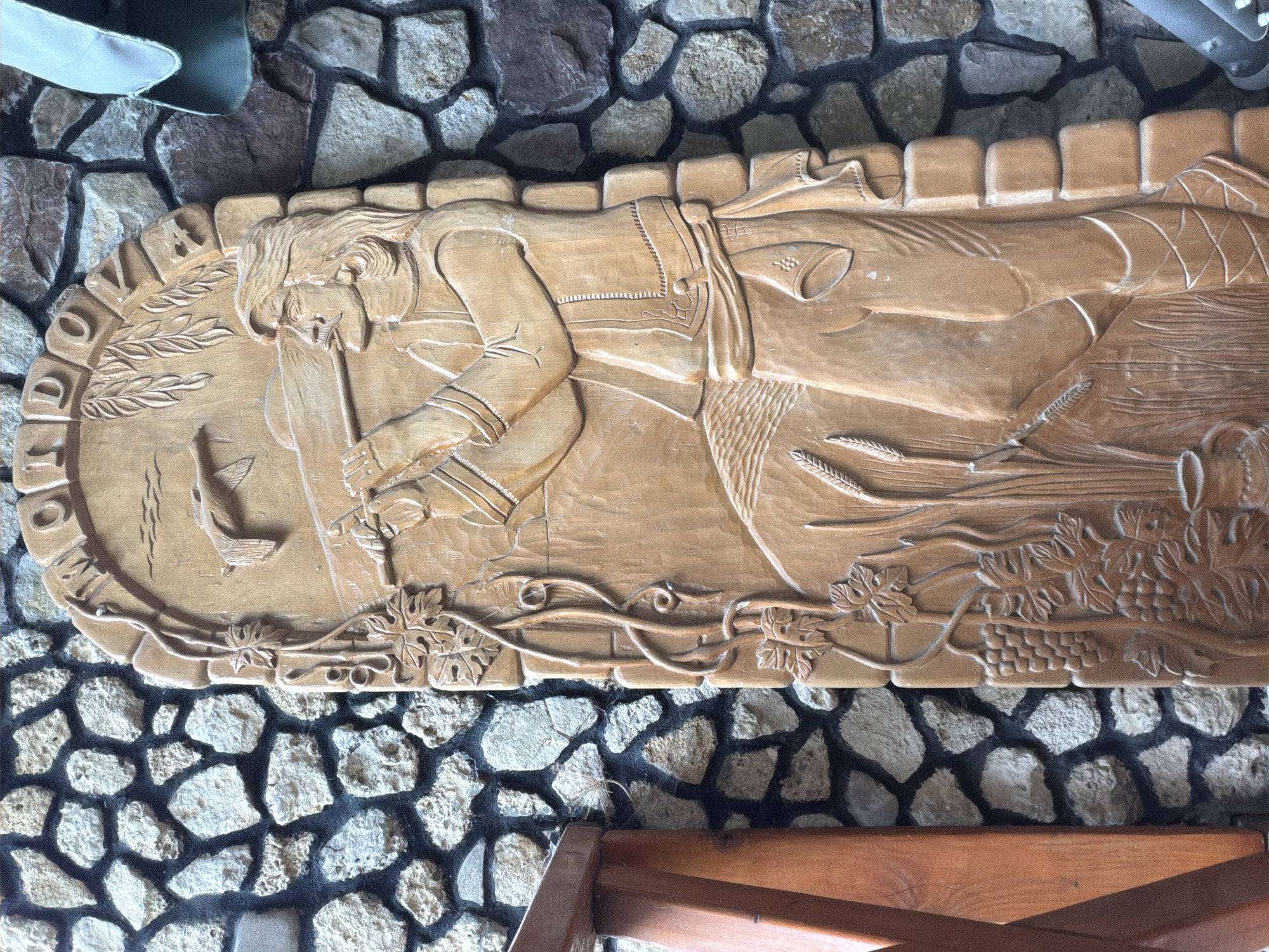 A carved wooden relief depicting a man playing a pipe is mounted on a stone wall. The carving shows a man with long hair and a mustache, wearing traditional clothing and standing amidst wheat stalks and grapevines. Above him, a bird is depicted in flight. The relief is framed by an arched top that contains carved letters which spell out "MONDROVA". The stone wall is made of irregular, rough-hewn rocks of various colors, including grey, beige, and brown, with dark mortar between them. A wooden railing is partially visible on the left side of the image. The overall setting appears to be outdoors, possibly a decorative element in the city of Oxentea, Moldova.