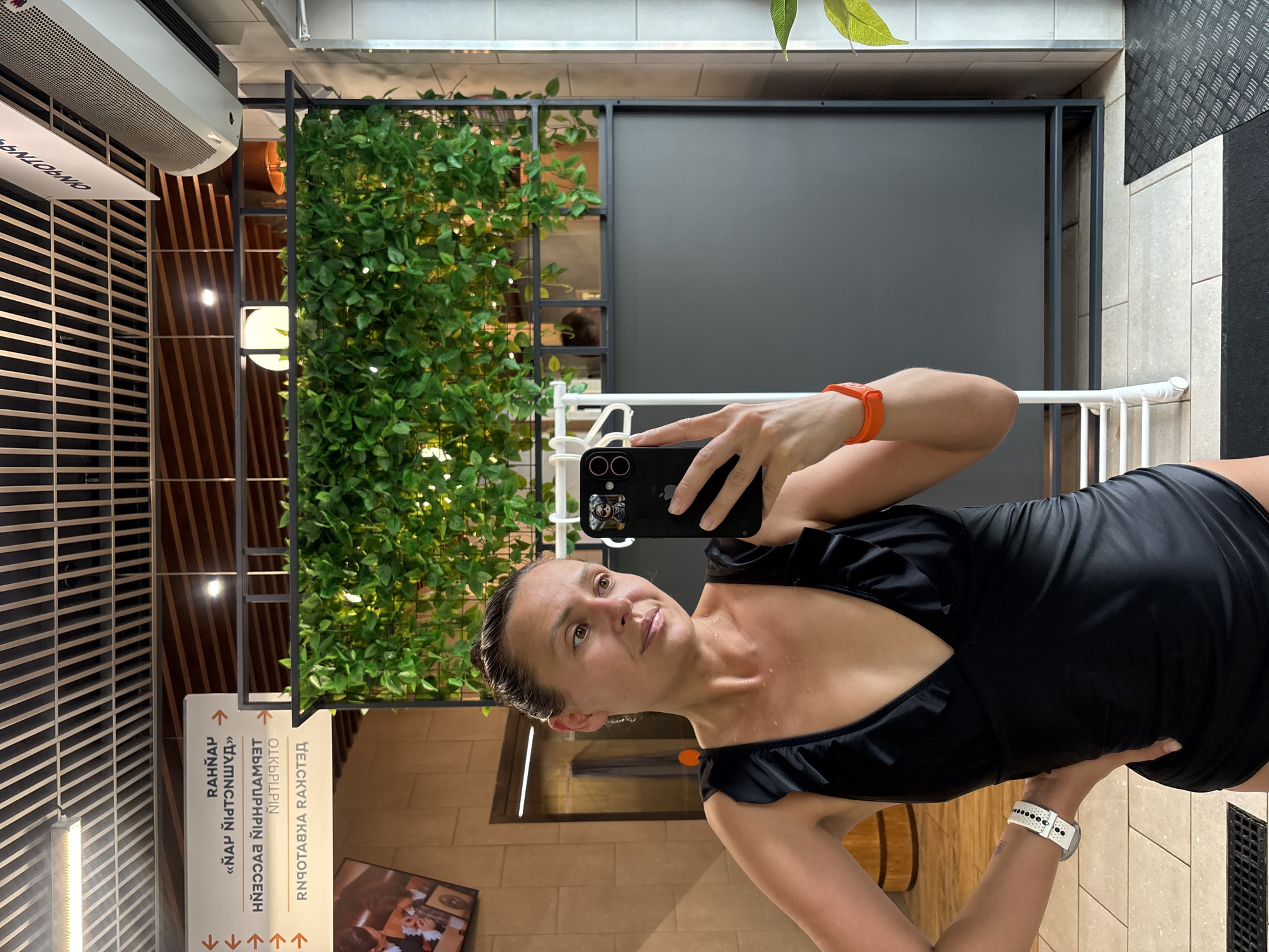 A woman with wet hair is taking a selfie in what appears to be a spa or wellness center. She is wearing a black bathing suit with ruffle details and an orange smartwatch on her left wrist and a white smartwatch on her right wrist. She is holding a black smartphone, likely an iPhone, in her right hand.
The background features a variety of elements including vertical wood slats, a sign with text in Russian, a leafy green plant wall, a gray wall panel, and tiled flooring.
Based on the Russian signage and the details, this location is likely in Votkinsk, Russia. The sign reads "РАННАР. «ПАР ЯЛТОНШУД» ТЕКШУМЯТ НИПМПАМЯТ NIPТLЯЯТО RNЯОТАВЯА ЯАЖОТЭД".