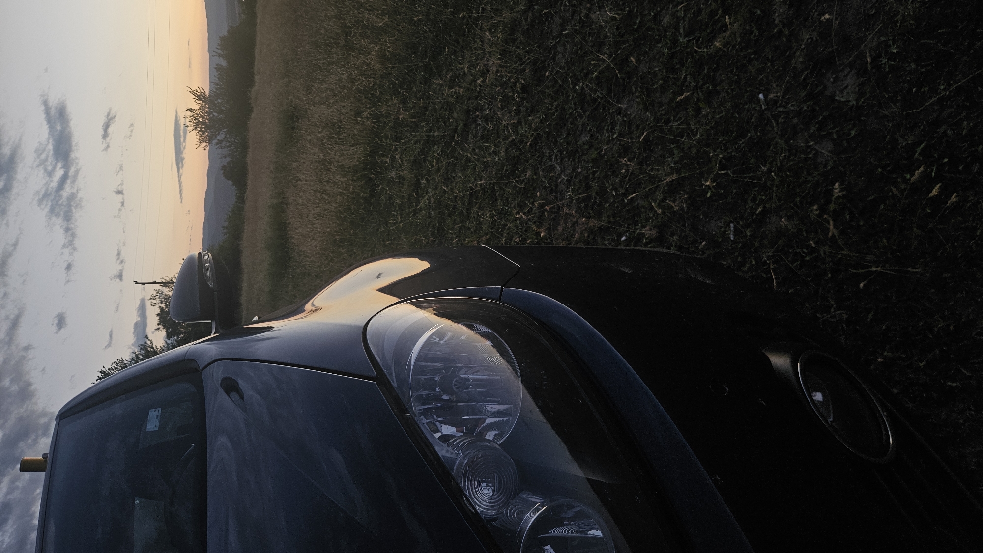 A low-angle, close-up shot features the front of a dark-colored car parked on grassy terrain during a sunset. The car's headlight, hood, and a portion of the windshield are visible on the left side of the frame. The warm tones of the sunset reflect off the car's paint. The background consists of rolling hills, distant trees, and a cloudy sky with hues of orange and purple. The right side of the image is dominated by dark, overgrown grass. No people are present in the scene. The overall setting suggests a rural or undeveloped area.