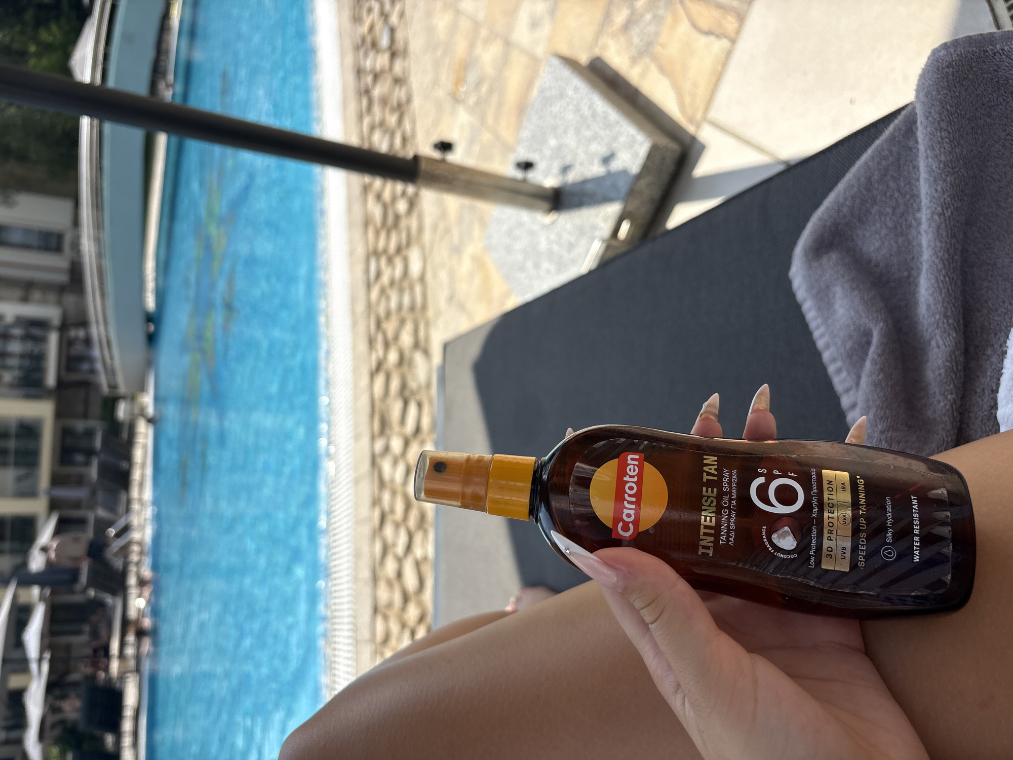 An outdoor scene features a person's hand holding a bottle of "Carroten Intense Tan Tanning Oil Spray SPF 6." The background shows a swimming pool with clear blue water, lounge chairs, umbrellas, and buildings. The lighting suggests a sunny day.