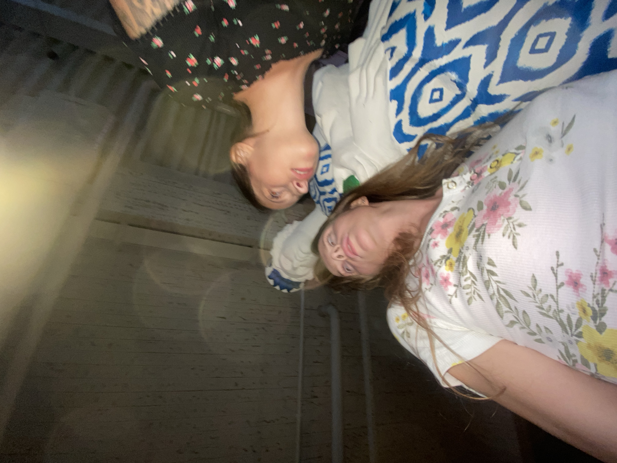 The image shows two women indoors. One woman, in the foreground and on the left, is lying down and looking upwards. She has long, light brown hair and is wearing a white t-shirt with a floral pattern of pink and yellow flowers and green leaves. The second woman is lying down to the right of the first woman and is looking towards the first woman. She has dark brown hair pulled back and is wearing a black t-shirt with a floral pattern of red flowers. Both women are on a surface covered with bedding that has a blue and white pattern. The background consists of a wooden ceiling or wall. The lighting suggests it is nighttime or a dimly lit indoor space. There are no specific city or location details discernible from the image itself that would place it in Solnechnoye, Russia.