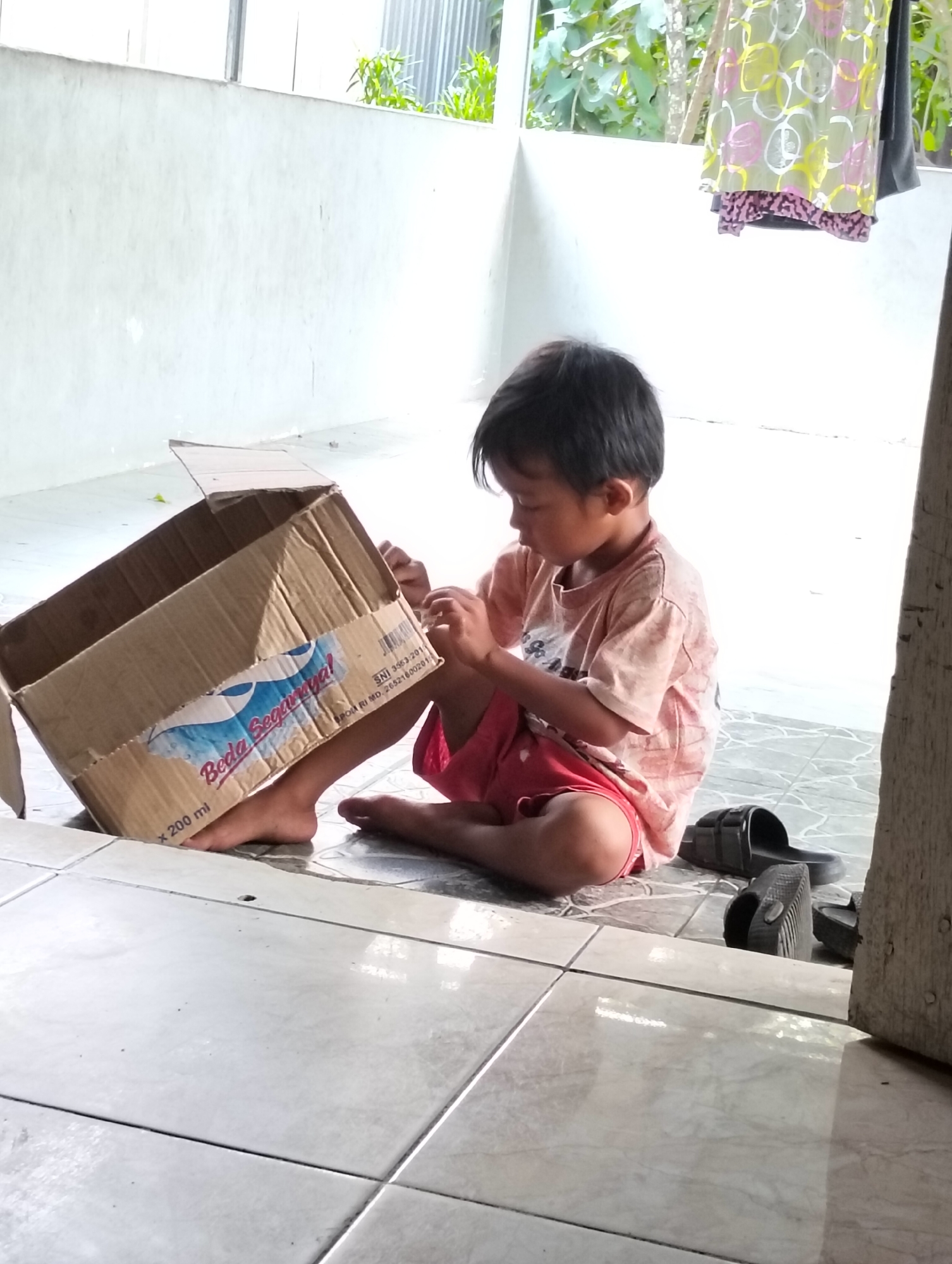 In Gambut, Indonesia, a young child sits on a tiled floor, engaged with a cardboard box. The child, wearing a light pink t-shirt with a subtle pattern and red shorts, has dark hair and is focused on the contents of the box. The cardboard box, partially open and tilted, displays a label with text and graphics, including "Beda Segarnya!" and "x 200 ml". A pair of black sandals rests on the floor near the child. The environment appears to be an outdoor or semi-outdoor space with a light-colored wall in the background, a section of corrugated metal, and some greenery visible. Clothes are also hanging on a line in the background. The scene suggests a moment of play or exploration by the child with the discarded packaging.