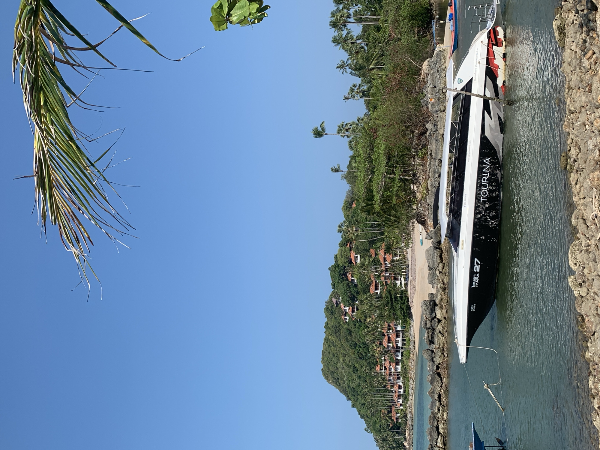The image depicts a coastal scene in Baan Tai, Thailand. A large, modern tour boat, labeled "TOURINA" with the number "27" and "IYARA" printed on its side, is docked at a stone-lined quay. The boat is primarily white with black and red accents. Clear turquoise water surrounds the boat. Behind the quay, a sandy beach stretches along the coast, backed by a steep, lush green hill covered in trees and dotted with buildings, potentially villas or resorts, with red-tiled roofs. Palm trees are interspersed throughout the landscape. The sky is clear and blue. A palm frond hangs in the upper left corner.