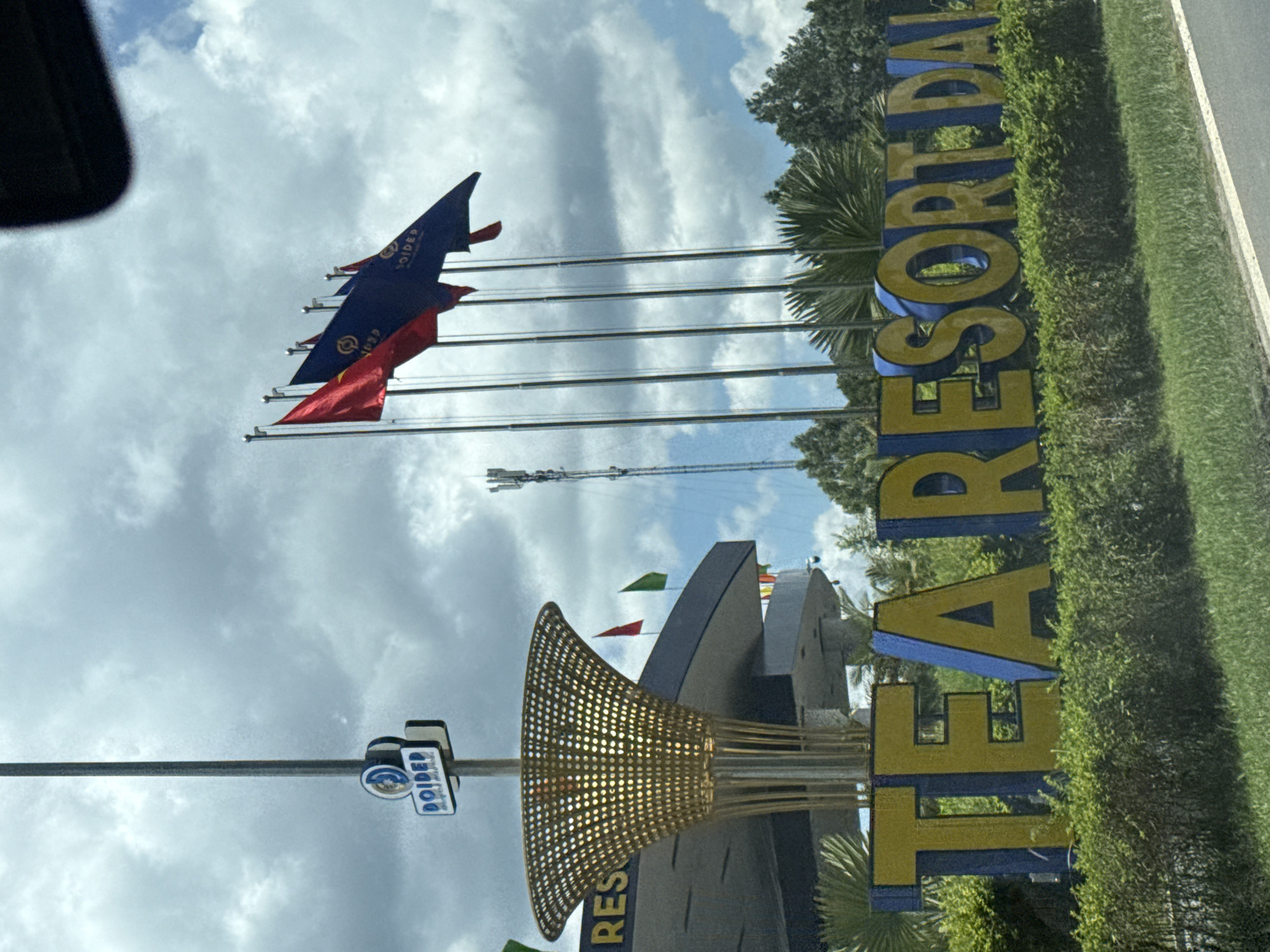 The image shows a large outdoor sign that reads "TEA RESORT DAI" in yellow and blue letters. The sign is set against a backdrop of green foliage and a cloudy sky. To the left of the sign, a tall pole with a circular blue and white logo bearing the word "DOIDEP" is visible. Several flags are flying on poles to the right of the sign, with some appearing to be red and blue. In the distance, a cellular tower can be seen. The scene appears to be a roadside view of an entrance to a resort. This location is Thôn 6, Vietnam.