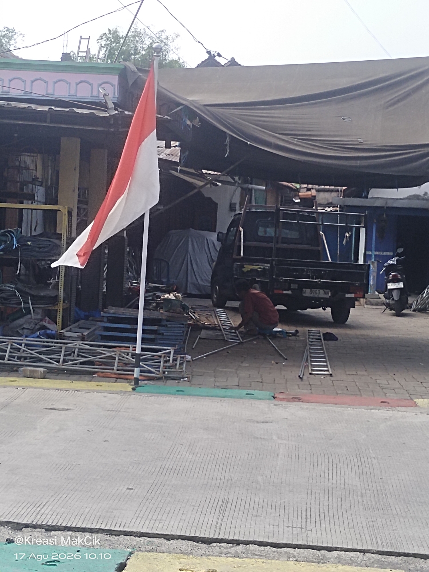 The image shows an outdoor scene in Sambi Bulu, Indonesia, with a focus on a pickup truck and a person working. The Indonesian flag, red and white, is prominently displayed on a flagpole to the left of the frame. In the center, a black pickup truck with an open bed is parked. To the left of the truck, a person wearing a red shirt is kneeling on the ground, assembling what appears to be a metal structure, possibly a ladder or scaffolding, with various metal components spread around them. Several ladders are visible, some assembled and some in parts. Behind the truck, a motorcycle is parked. The environment appears to be a street or an open lot with a mix of concrete and brick paving. Buildings with awnings and corrugated metal roofs are visible in the background, suggesting a commercial or workshop area. The overall impression is of a scene of ongoing work or setup in a bustling Indonesian locale.