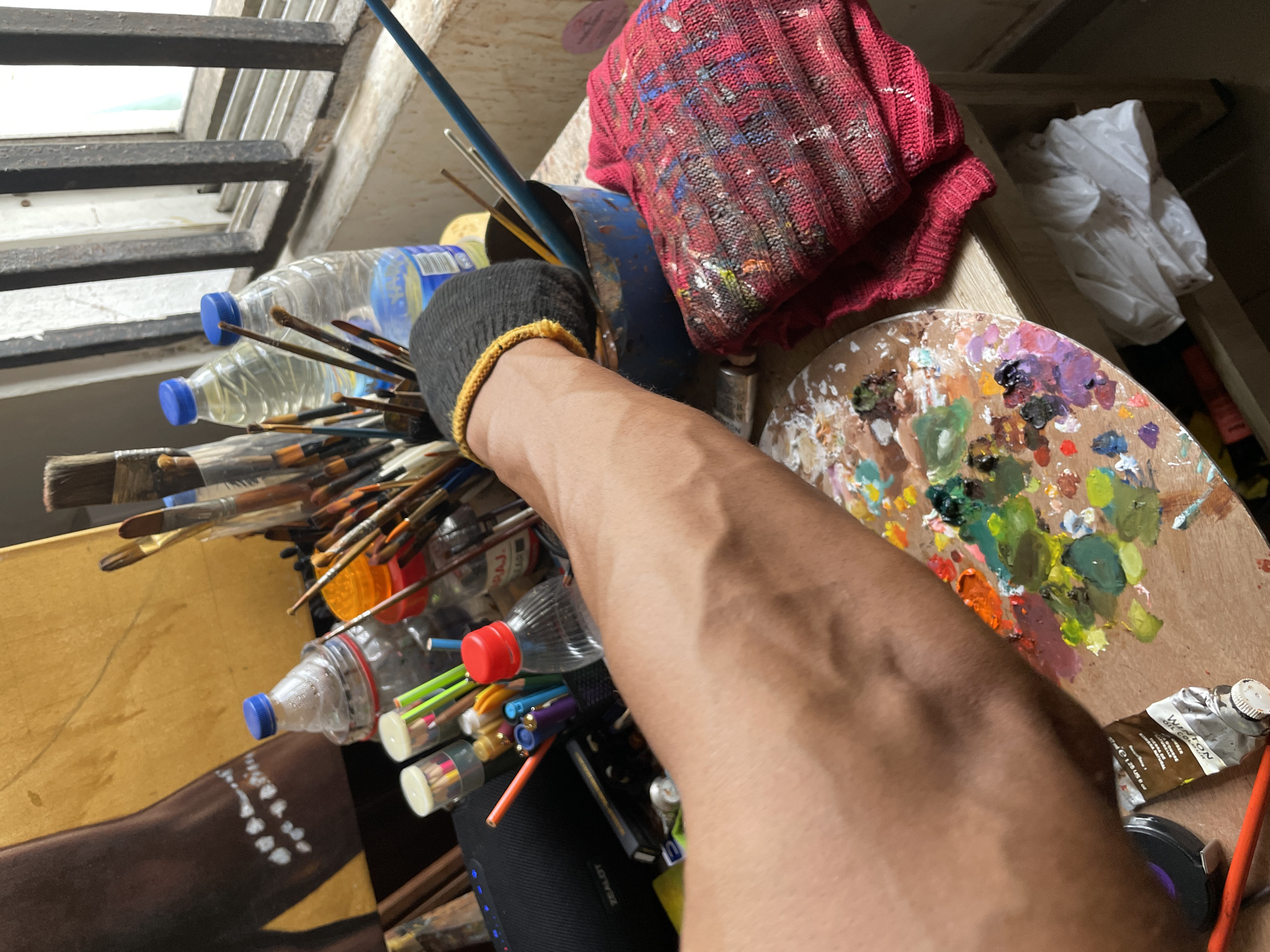 The image shows an artist's workspace, likely indoors in Ado, Nigeria. A collection of paintbrushes, some stored in recycled plastic water bottles, is visible. A paint-stained red cloth rests on a blue container. A wooden palette covered in assorted paints is nearby. A visible arm wearing a fingerless glove reaches into the brush container. Part of a painting is visible, showing a figure with beaded jewelry. A tube of Wilton oil paint is also present. The environment appears cluttered and artistic.