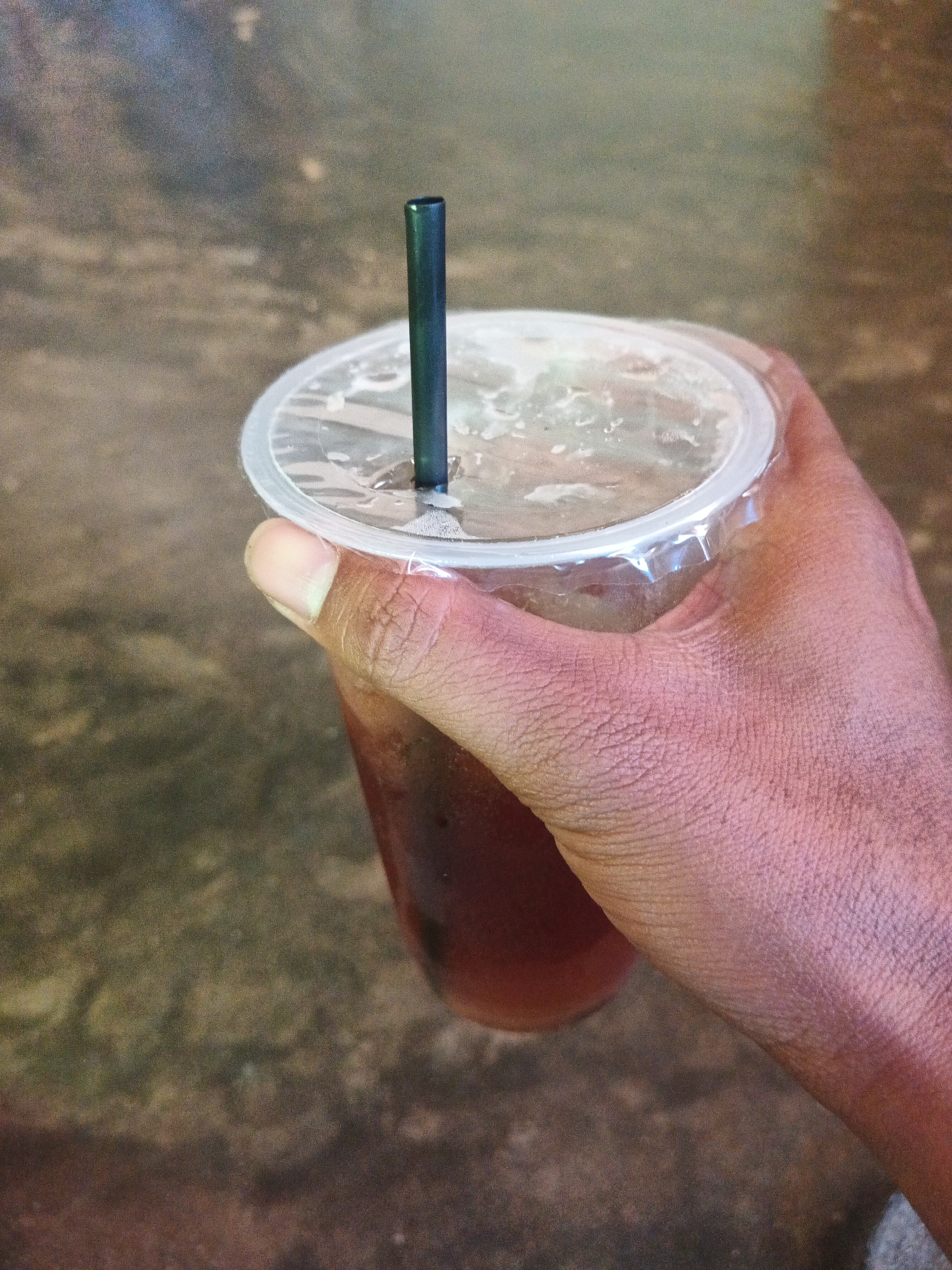 A close-up, eye-level shot shows a person's hand holding a clear plastic cup filled with a dark liquid and ice. A black plastic straw is inserted through the lid. The background is a blurred, textured surface that appears to be concrete or asphalt, possibly a sidewalk or outdoor seating area. The lighting is subdued, casting subtle shadows and highlights on the hand and cup. The image is taken from a high angle looking down into the cup. The overall impression is one of casual consumption, possibly in an urban or cafe setting. No specific landmarks or identifiable features place this image in any particular city or location in Indonesia.