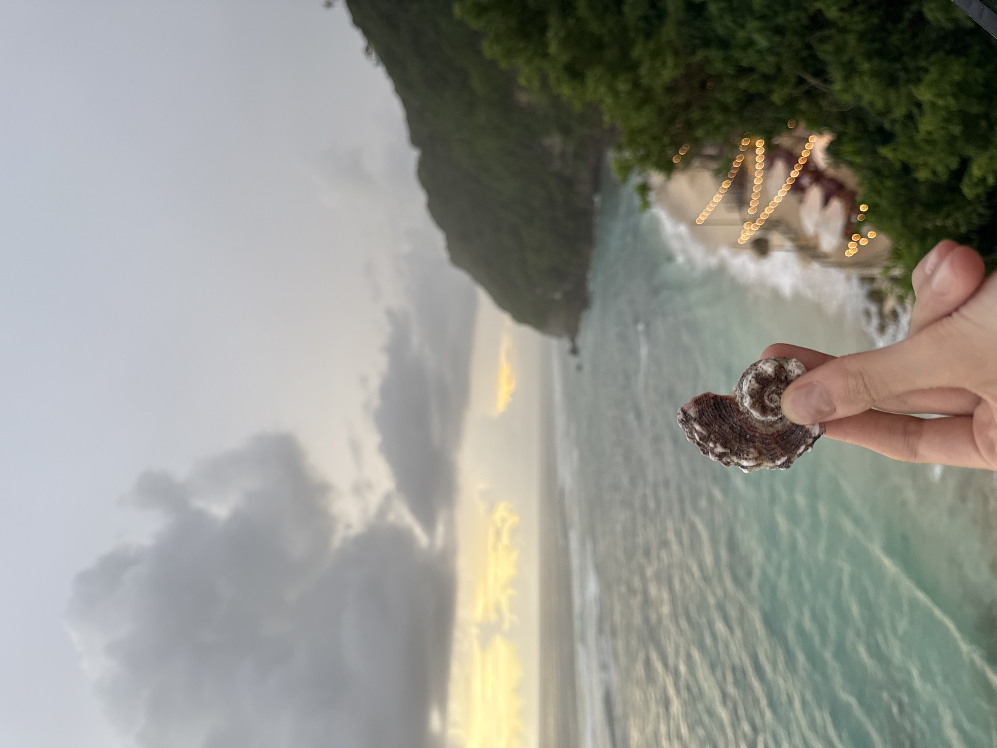 The image shows a person holding a seashell in their fingers. In the background, an aerial view of the coast shows a beach with waves crashing on the shore, a cliff covered in lush green vegetation, and a section of the sky with clouds. String lights are visible on the beach. The location is Kuta Selatan, Indonesia.